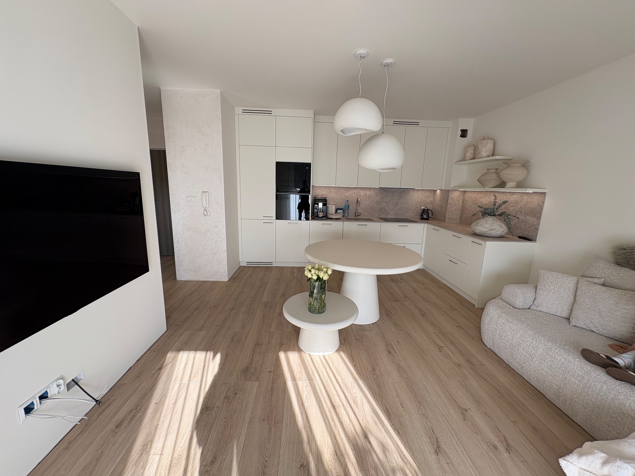 Jasne, minimalistyczne wnętrze: biała kuchnia z marmurowym blatem, okrągły stół i sofa. Uwagę przykuwają designerskie lampy i drewniana podłoga z promieniami słońca.