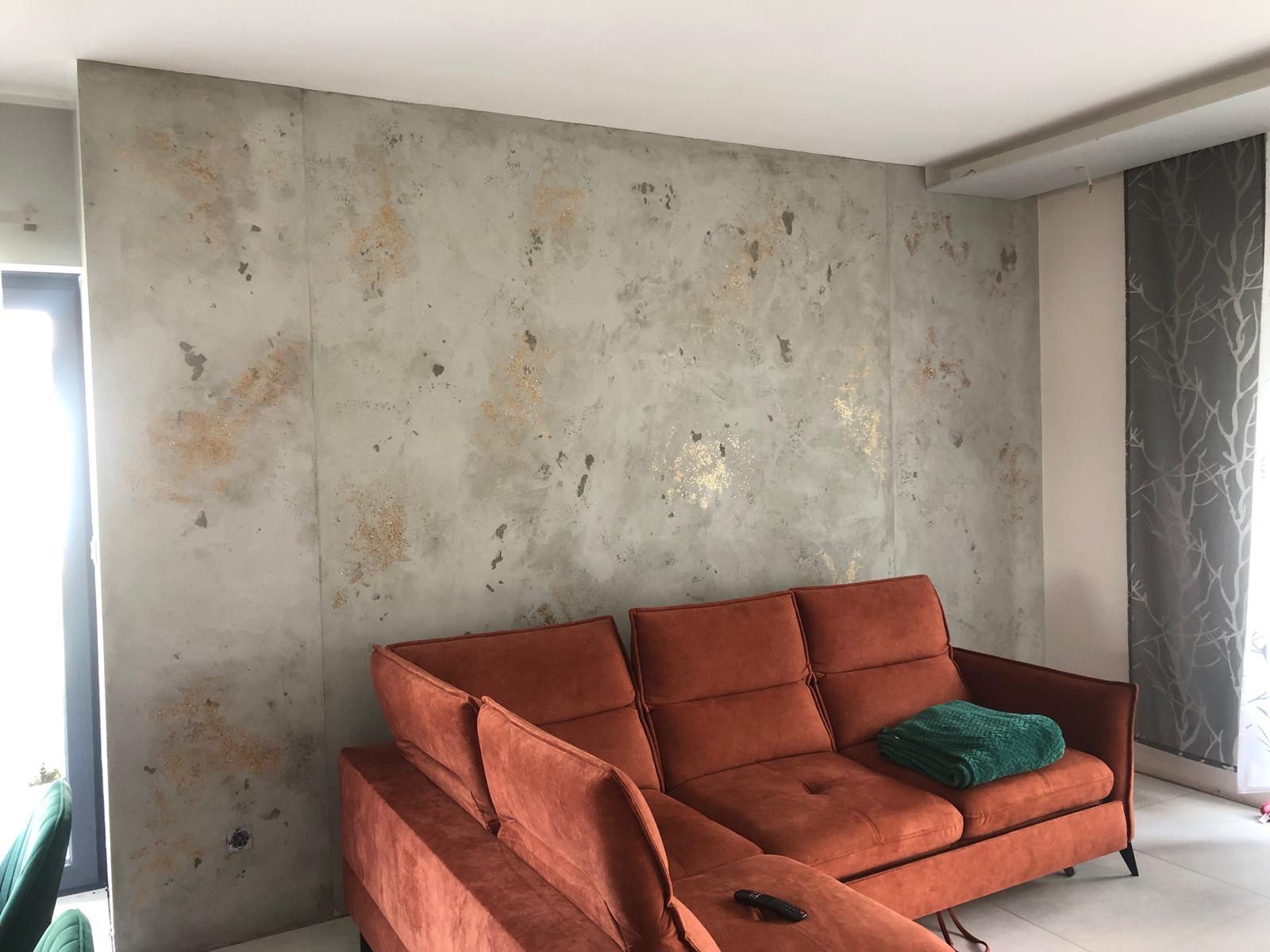 Ściana w salonie z dekoracyjnym tynkiem imitującym beton, z dodatkiem złotych akcentów. Pomarańczowa sofa z zielonym pledem. Nowoczesne wykończenie wnętrza.