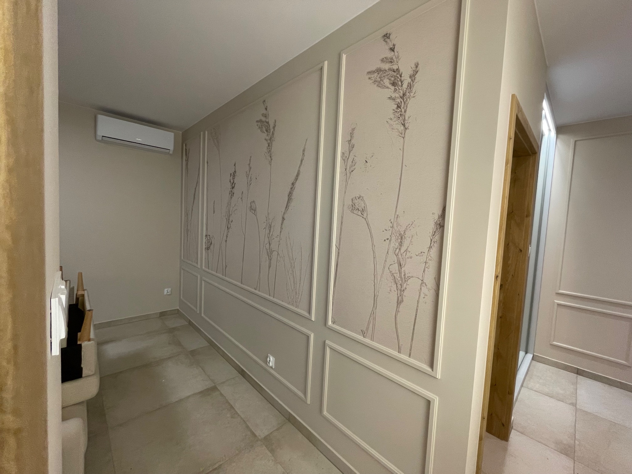 Eleganckie wykończenie wnętrza: ściana z dekoracyjnymi panelami i motywem roślinnym, jasna podłoga, klimatyzator. Widok na sąsiednie pomieszczenie.