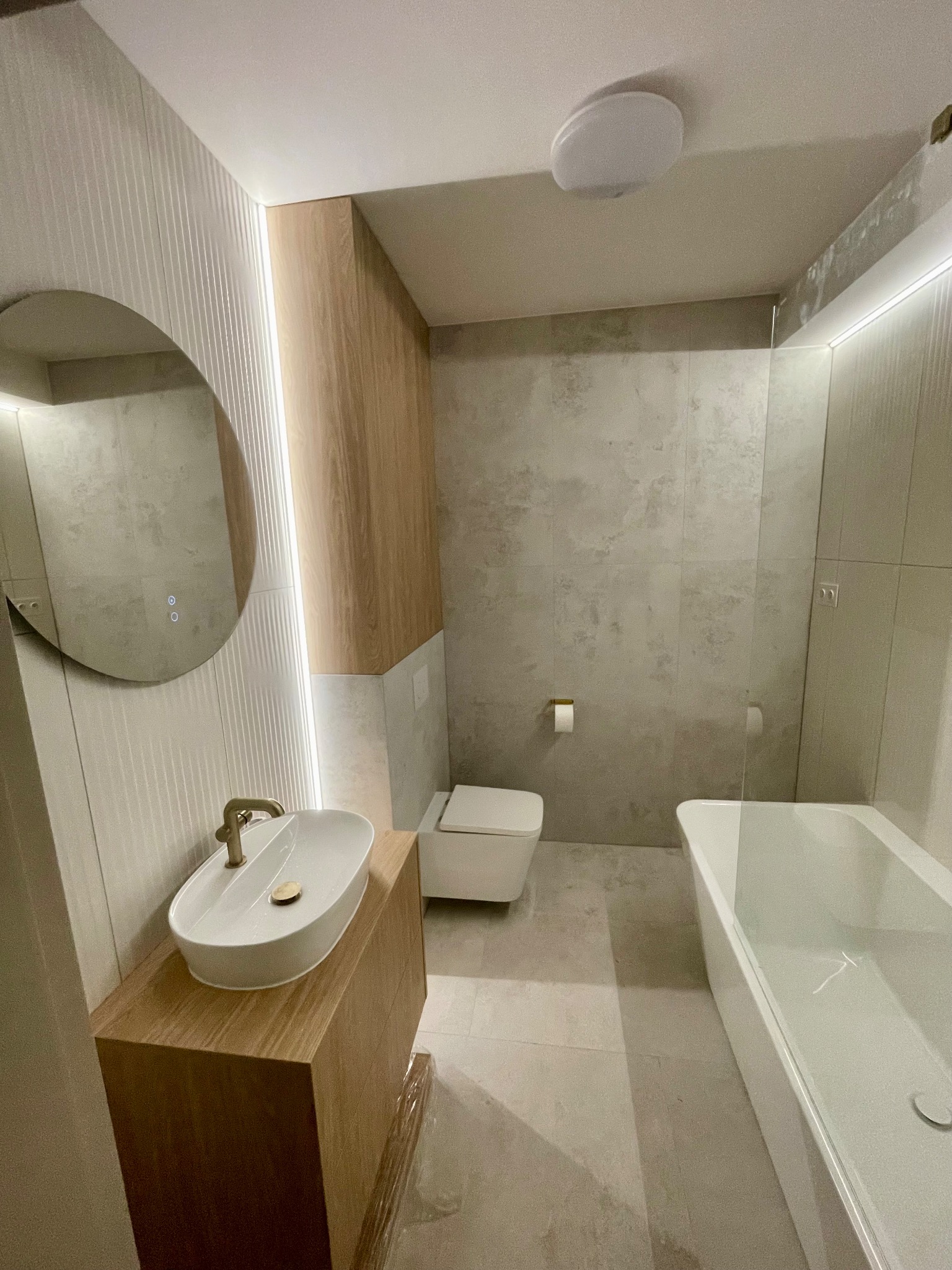 Nowoczesna łazienka z owalną umywalką na drewnianej szafce, podświetlanym lustrem i wanną za szklaną ścianką. Stonowana kolorystyka płytek i oświetlenie LED.