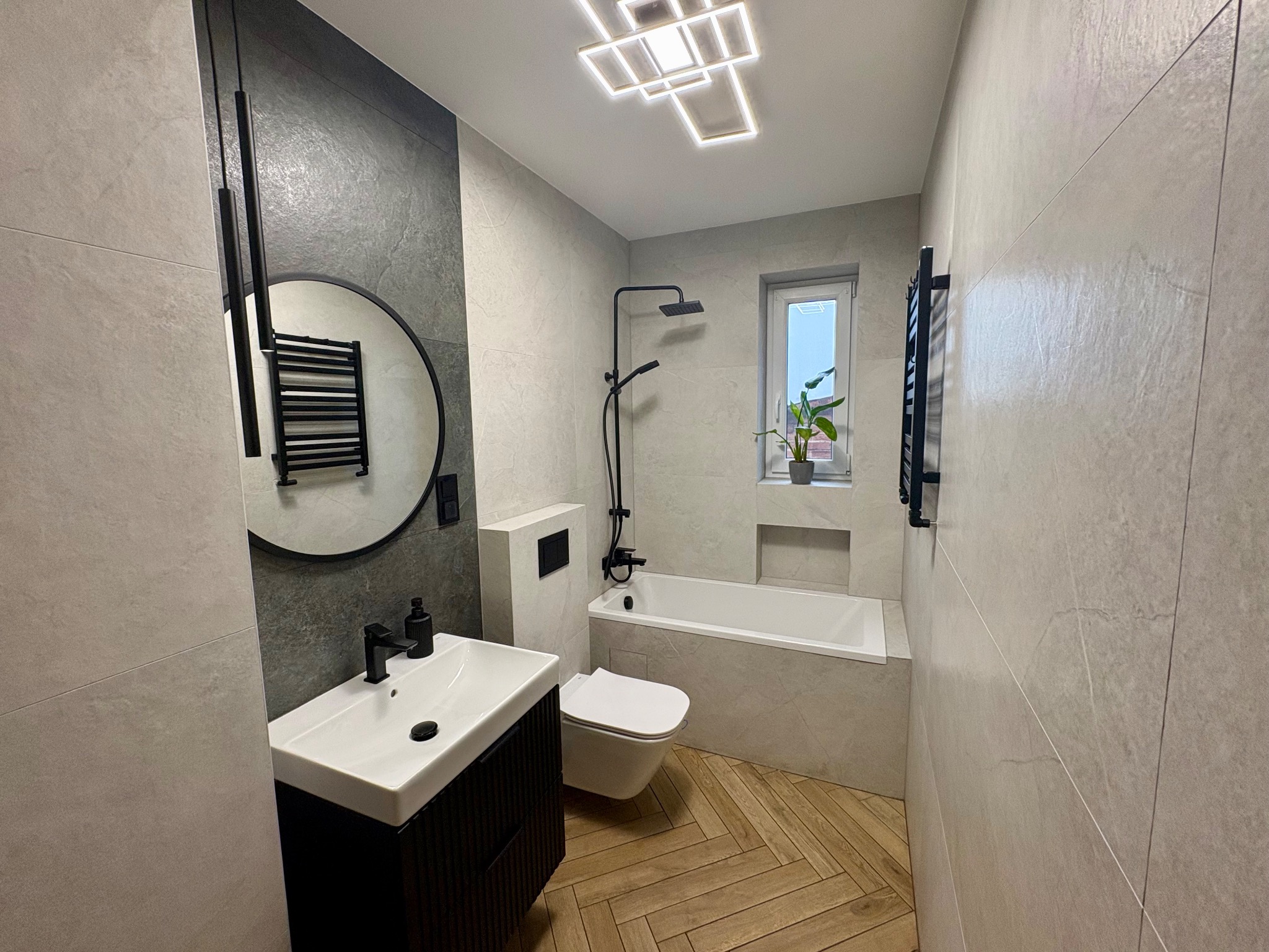 Nowoczesna łazienka z szarymi płytkami, drewnianą podłogą w jodełkę i czarnymi akcentami: armatura, oświetlenie, okrągłe lustro z grzejnikiem.