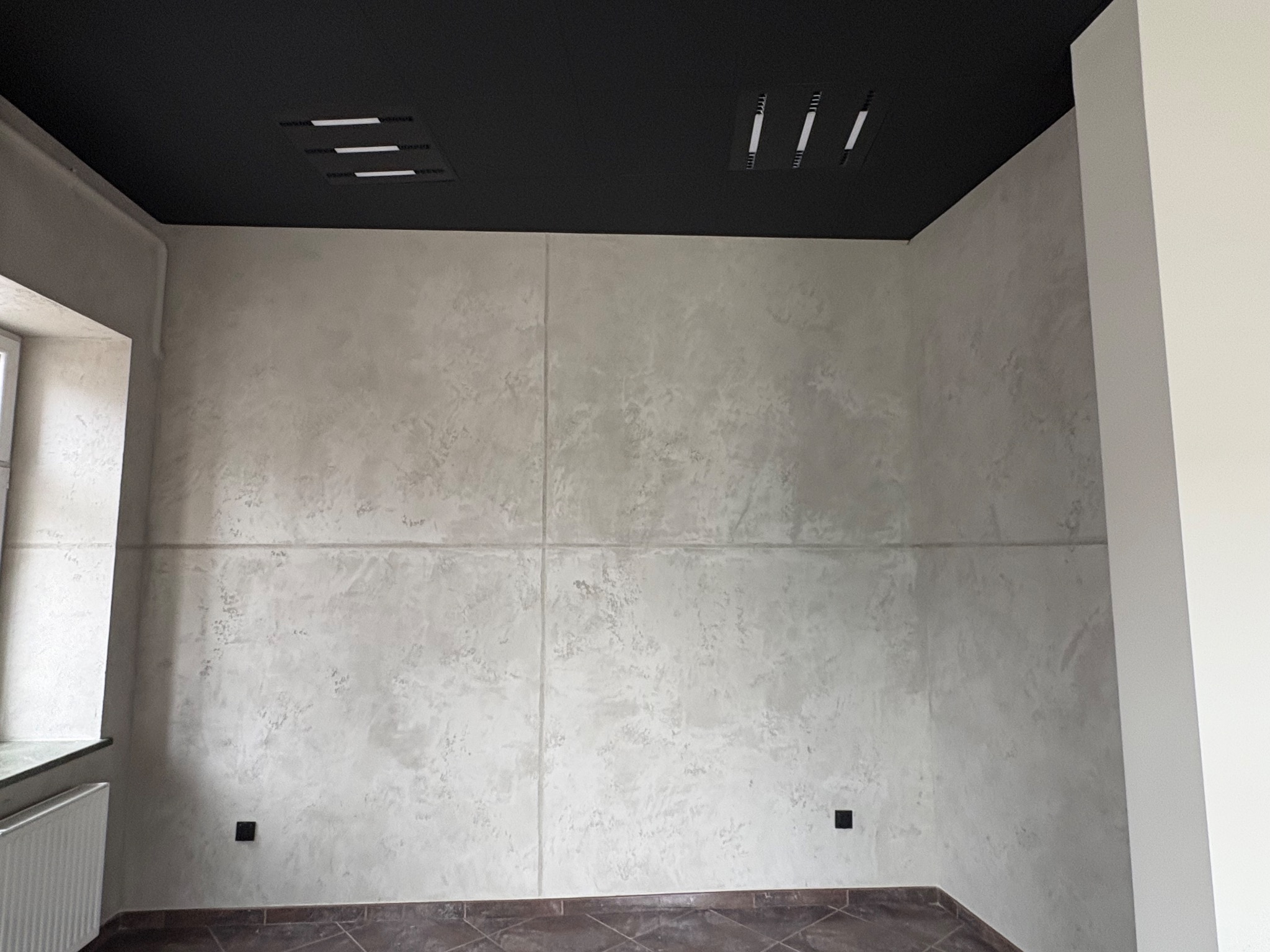 Wykończone wnętrze: ściana z efektem dekoracyjnym w odcieniach szarości, czarny sufit z oświetleniem, brązowa podłoga z płytek. Widok na fragment okna i grzejnik.