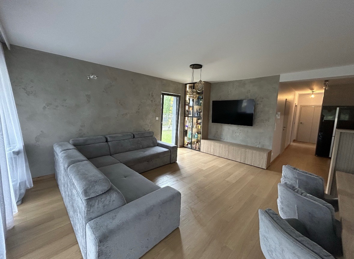 Szary narożnik w salonie z efektem betonu na ścianie, drewniana podłoga i szafka RTV. Widoczna przeszklona witryna z oświetleniem i telewizor. Nowoczesne wykończenie.