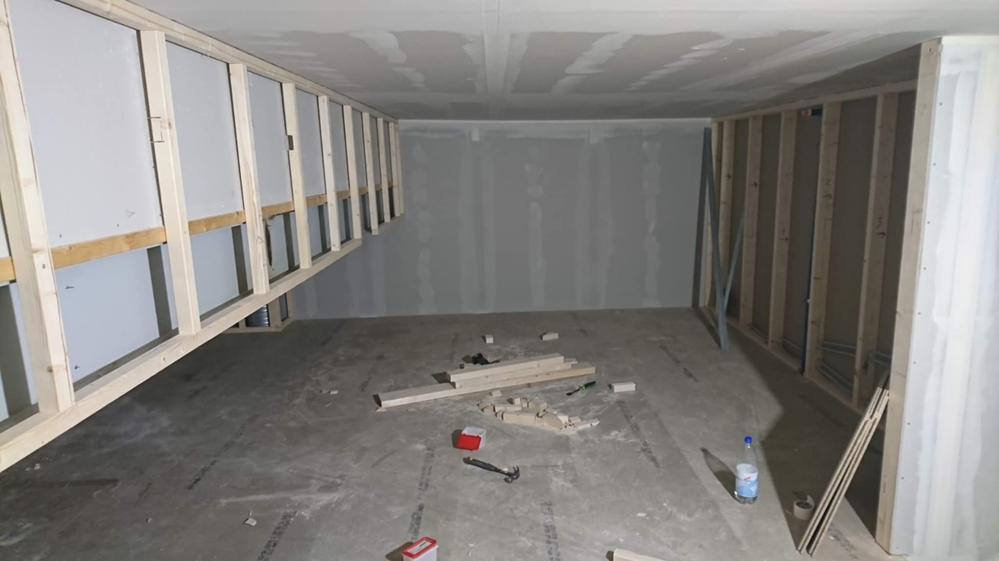 Wnętrze w trakcie prac z płytami gipsowo-kartonowymi: widoczne drewniane stelaże, częściowo zamontowane płyty, narzędzia i materiały budowlane na surowej podłodze.