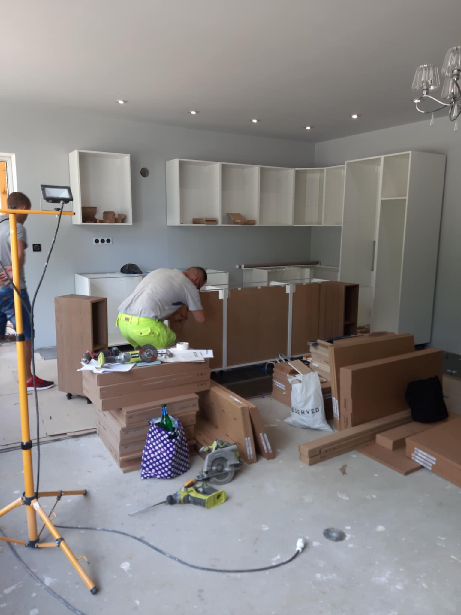 Montaż białych szafek kuchennych w surowym wnętrzu. Widoczne narzędzia, kartony i oświetlenie robocze. Jeden z monterów w trakcie pracy.