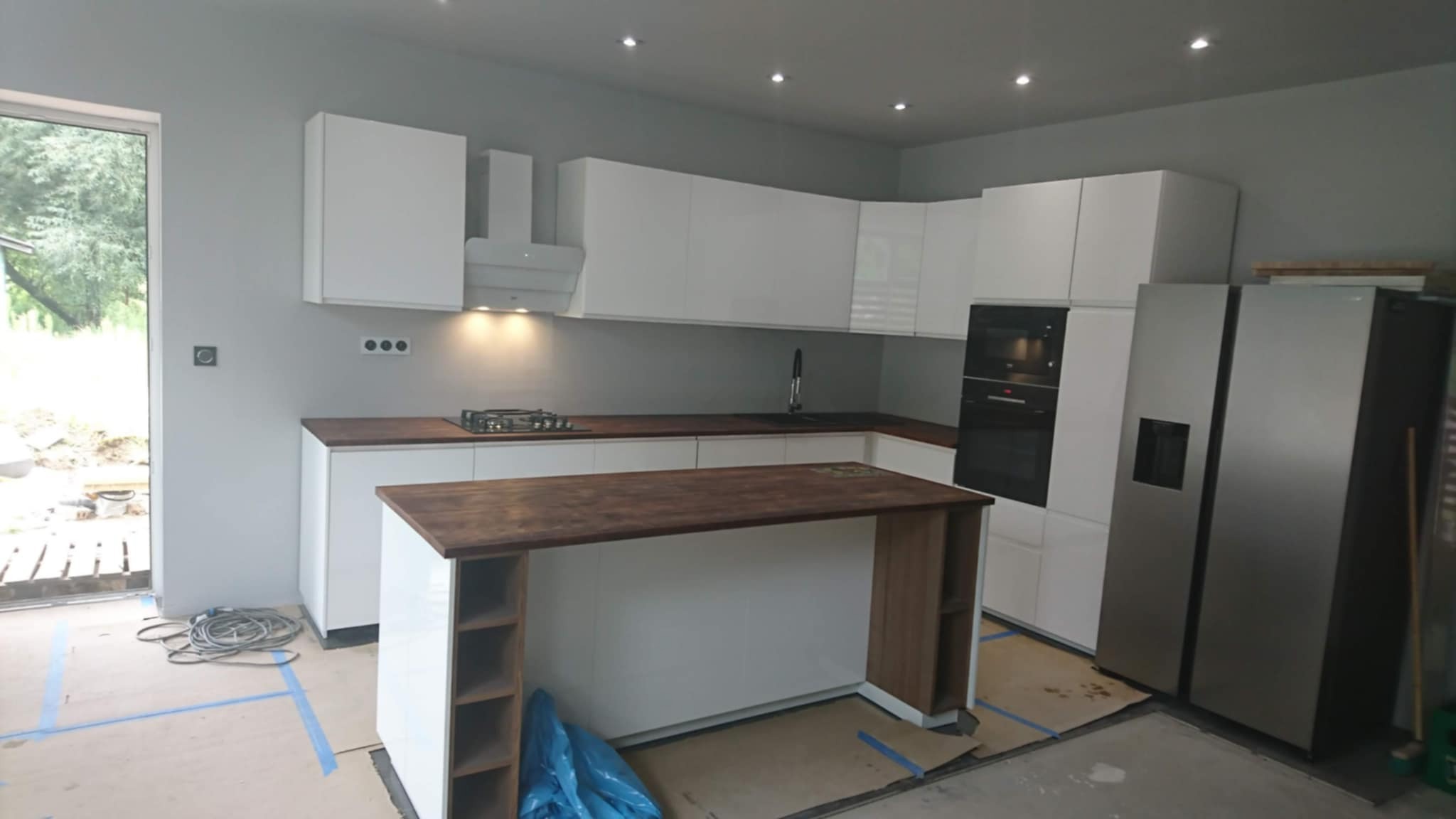 Nowoczesna kuchnia z białymi szafkami i drewnianym blatem, wyspą. Widoczne sprzęty AGD i okno. Podłoga zabezpieczona kartonem i taśmą malarską.