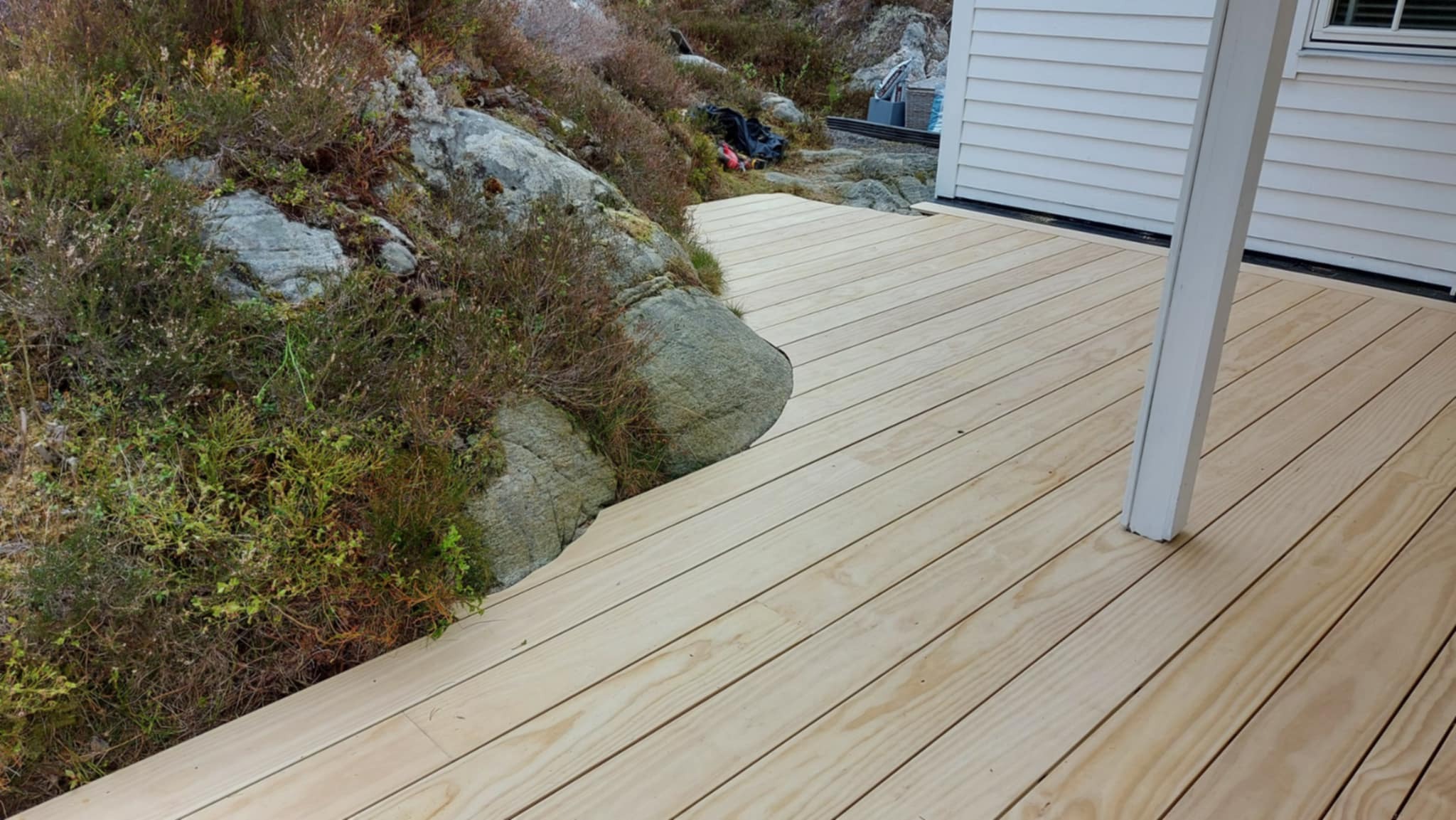 Jasna, drewniana podłoga tarasu przylegająca do skały i zieleni, z fragmentem białej ściany budynku w tle. Widok z góry, podkreślający naturalne materiały.