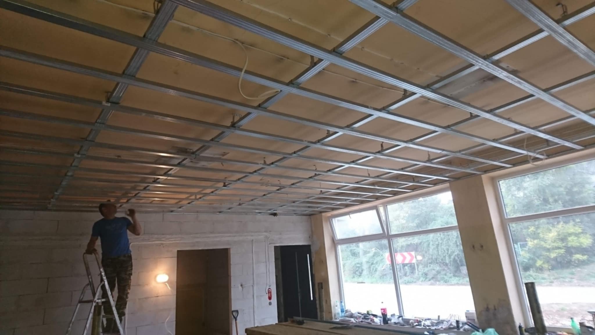 Montaż sufitu podwieszanego z płyt karton-gips: widoczna metalowa konstrukcja i częściowo zamontowane płyty. Pracownik na drabinie w trakcie pracy. Duże okna w tle.
