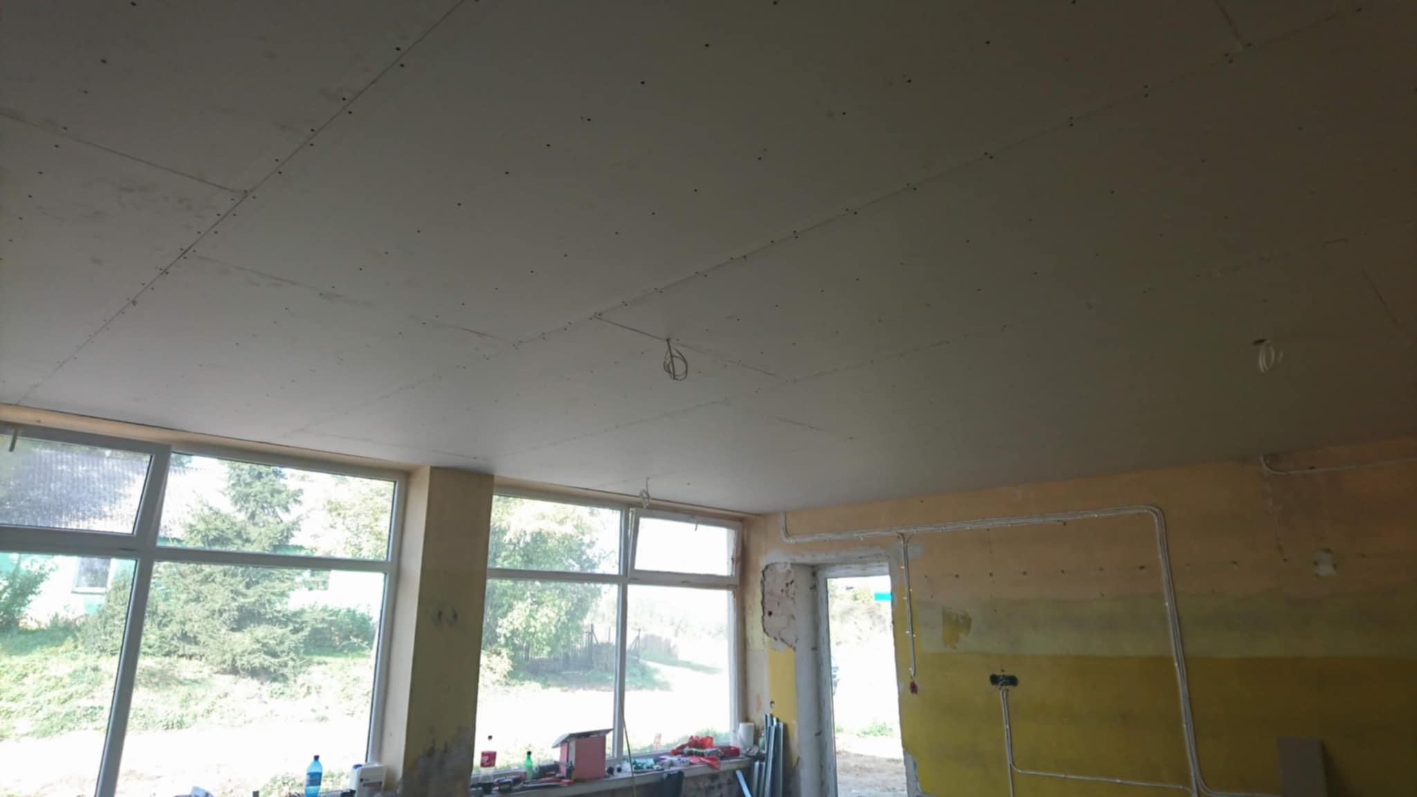 Sufit z płyt kartonowo-gipsowych w trakcie montażu, widoczne łączenia i wkręty. W tle okno z widokiem na zieleń i fragment ściany z instalacją elektryczną.