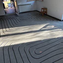 Jarosław Jacek - Szare rury ogrzewania podłogowego ułożone spiralnie na podłodze w jasnym pomieszczeniu z oknem i grzejnikiem. Widoczny fragment drzwi i osoba w tle.