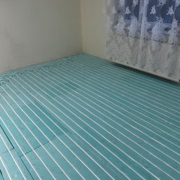 Jarosław Jacek - Błękitna izolacja podłogowa z białymi rurami ogrzewania podłogowego, ułożona w regularnych odstępach w rogu pokoju z oknem przysłoniętym białą, kwiecistą firanką.