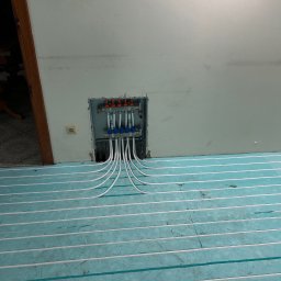 Jarosław Jacek - Błękitna izolacja podłogowa z białymi rurami ogrzewania podłogowego, w tle rozdzielacz w ścianie. Widok z góry, ukończony etap instalacji.