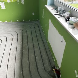 Jarosław Jacek - Szare rury ogrzewania podłogowego ułożone w jastrychu, widoczne fragmenty zielonej ściany z niedomalowkami i okno z narzędziami na parapecie.