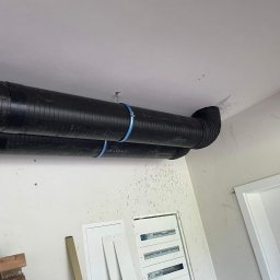 Jarosław Jacek - Dwa czarne przewody wentylacyjne przymocowane do sufitu za pomocą niebieskich obejm, widoczne skrzynki elektryczne na ścianie i fragment ościeżnicy drzwi.