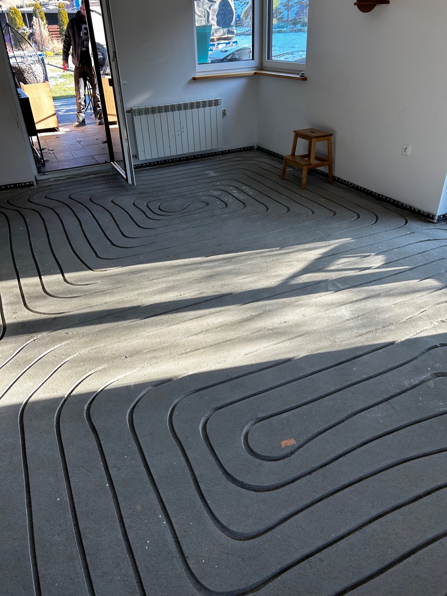 Szare rury ogrzewania podłogowego ułożone spiralnie na podłodze w jasnym pomieszczeniu z oknem i grzejnikiem. Widoczny fragment drzwi i osoba w tle.