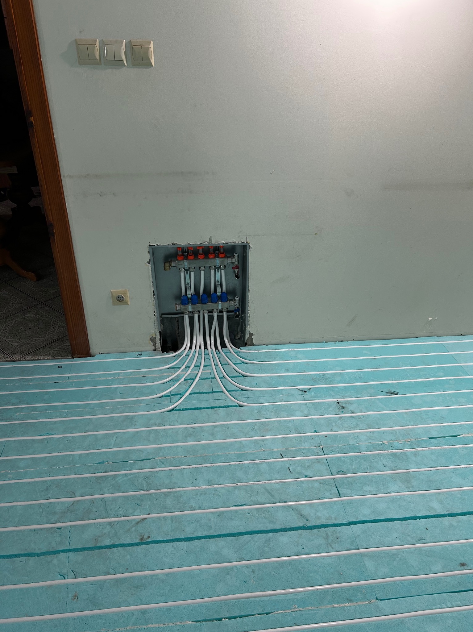 Błękitna izolacja podłogowa z białymi rurami ogrzewania podłogowego, w tle rozdzielacz w ścianie. Widok z góry, ukończony etap instalacji.