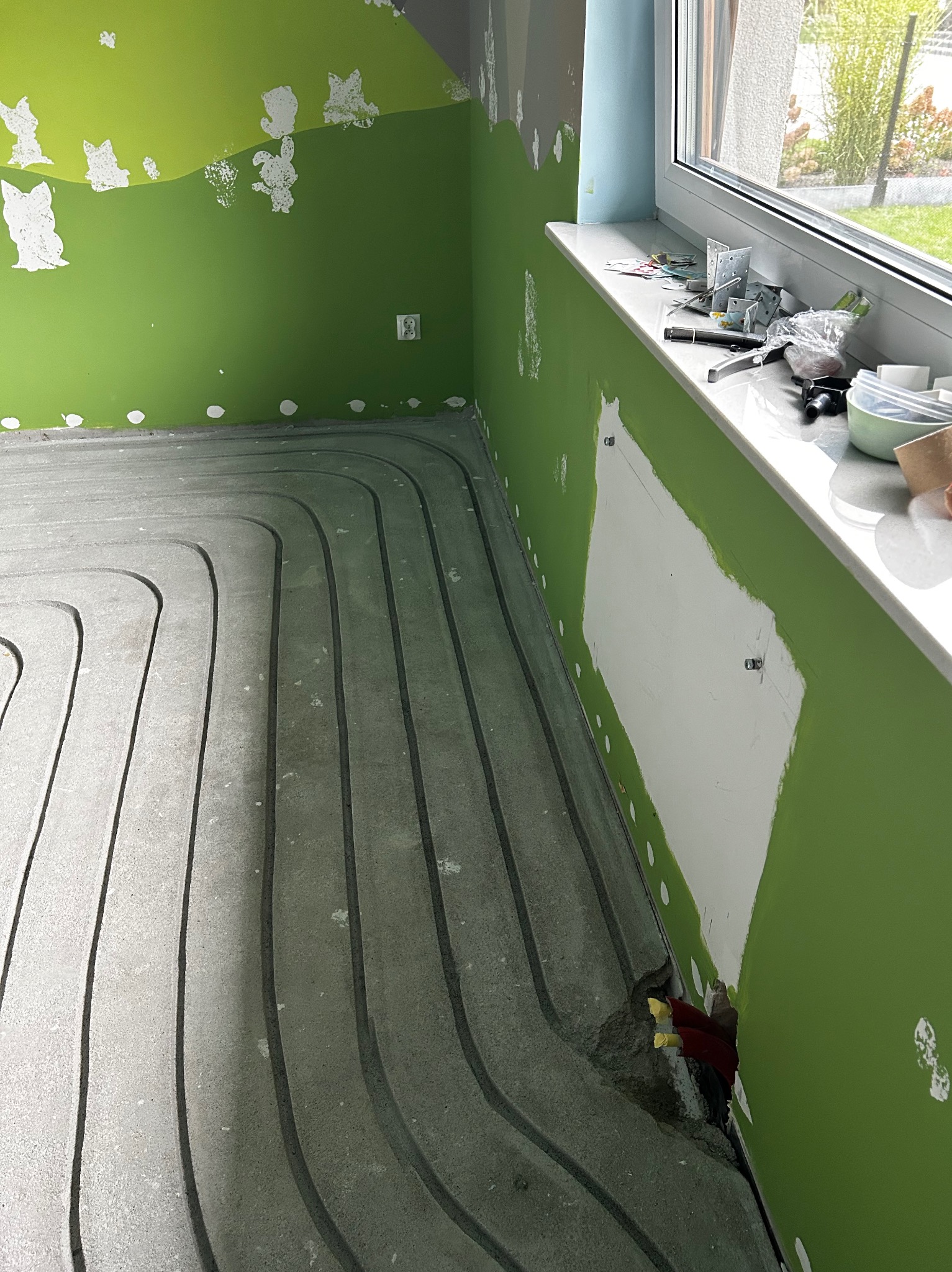 Szare rury ogrzewania podłogowego ułożone w jastrychu, widoczne fragmenty zielonej ściany z niedomalowkami i okno z narzędziami na parapecie.