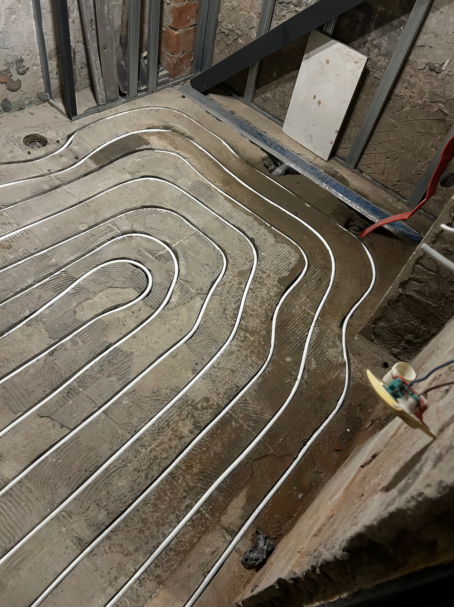 Białe rurki ogrzewania podłogowego ułożone spiralnie na szarej, nierównej powierzchni. Widoczne elementy konstrukcyjne i fragment ściany z cegły.