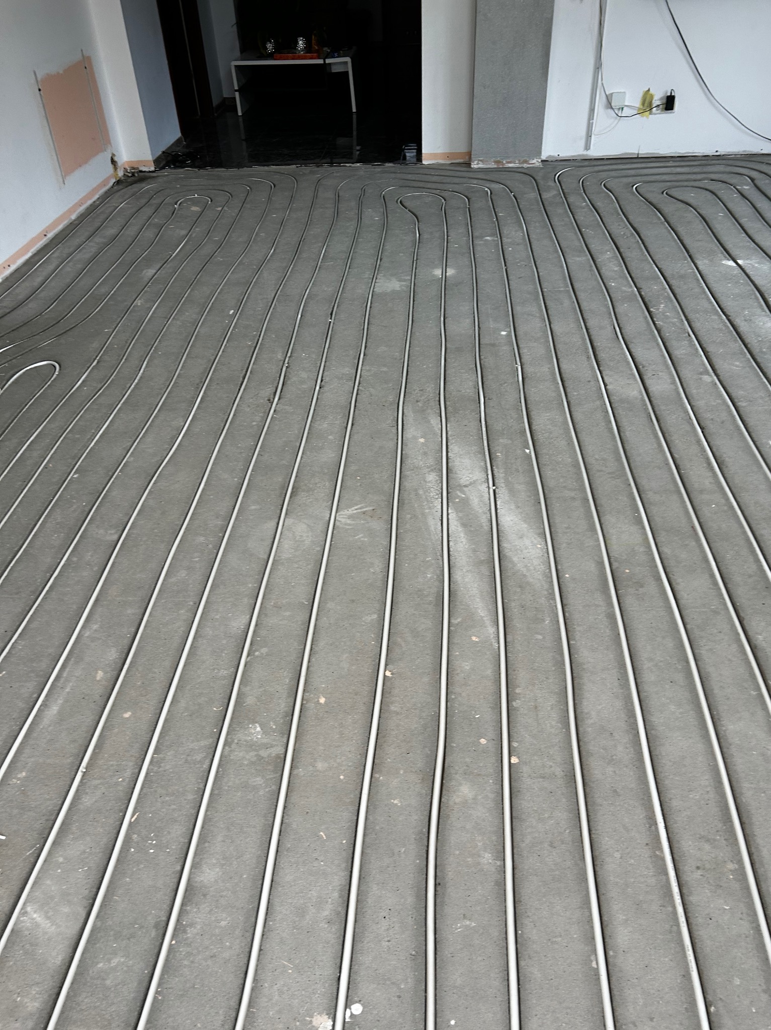 Szare rury ogrzewania podłogowego ułożone równolegle na betonowej wylewce, widok z góry, z fragmentem instalacji elektrycznej przy ścianie w tle.