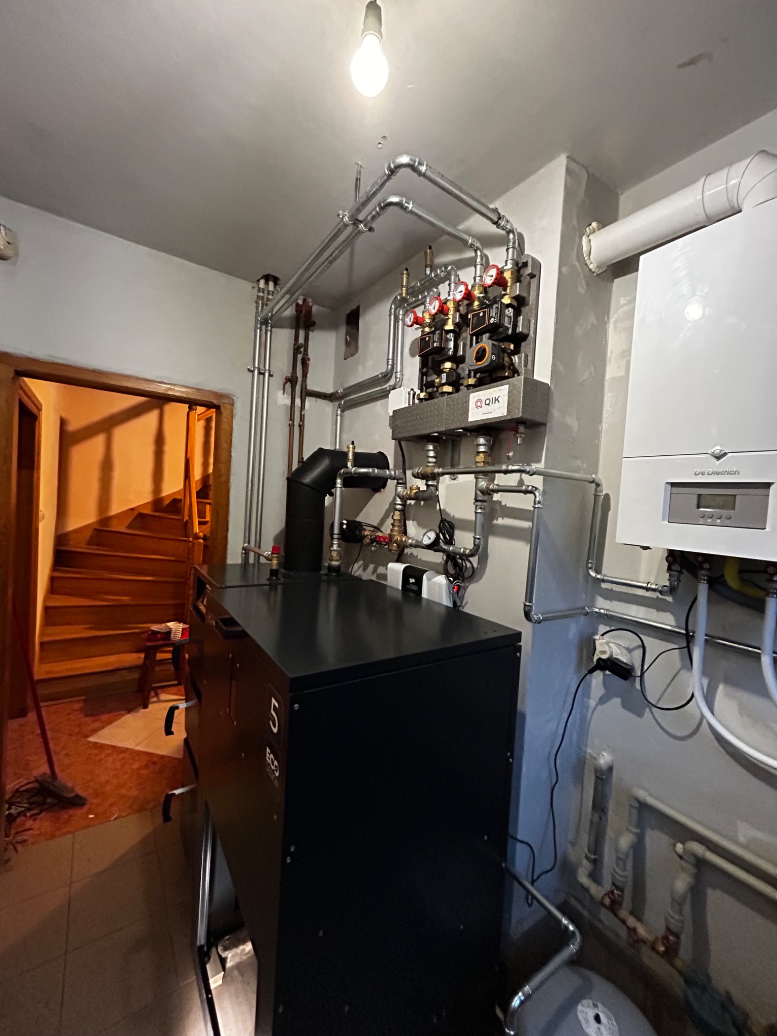 Czarny piec CO z podłączeniami, widoczne rury, zawory i biały bojler. Instalacja w kotłowni z oświetleniem żarówkowym i schodami w tle.