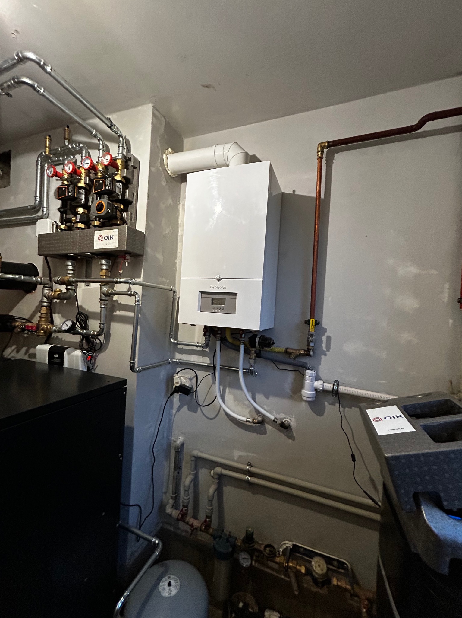 Biały piec gazowy De Dietrich z instalacją hydrauliczną i rozdzielaczem w Chorzelowie. Widoczne rury miedziane i zawory. Szara ściana w tle.