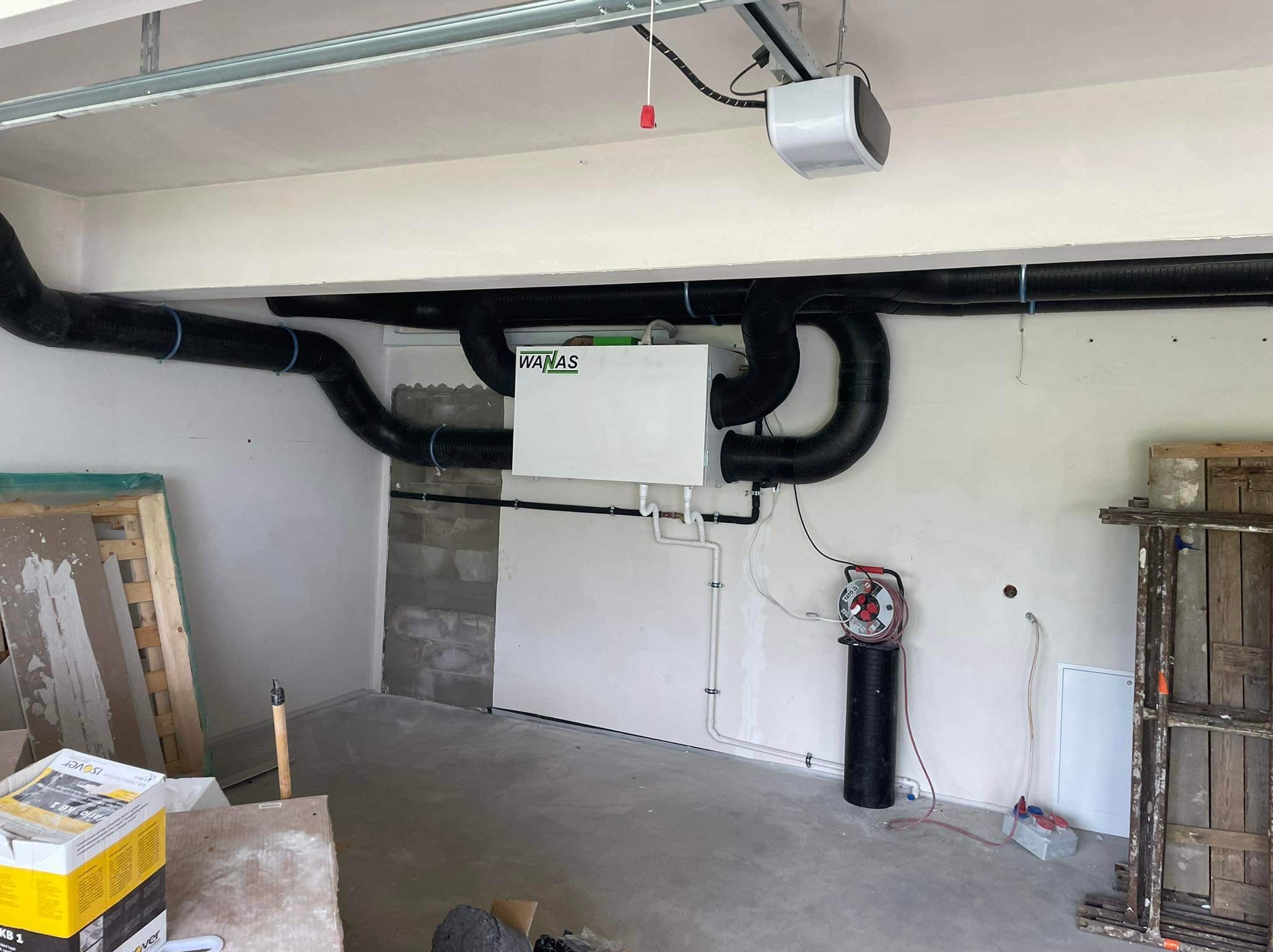 Biała centrala rekuperacyjna WANAS z czarnymi rurami wentylacyjnymi na ścianie w surowym pomieszczeniu. Widoczne narzędzia i materiały budowlane.