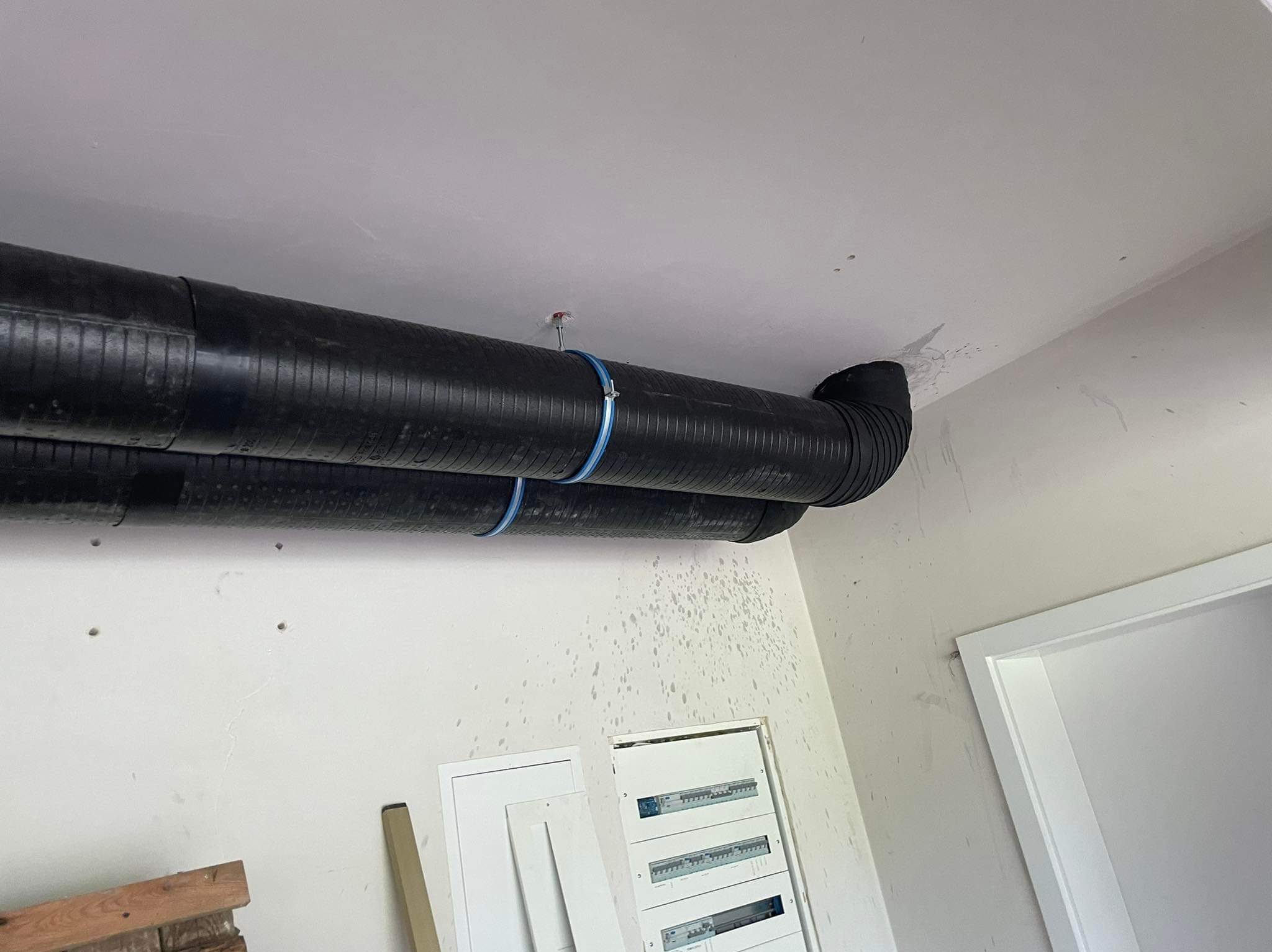 Dwa czarne przewody wentylacyjne przymocowane do sufitu za pomocą niebieskich obejm, widoczne skrzynki elektryczne na ścianie i fragment ościeżnicy drzwi.