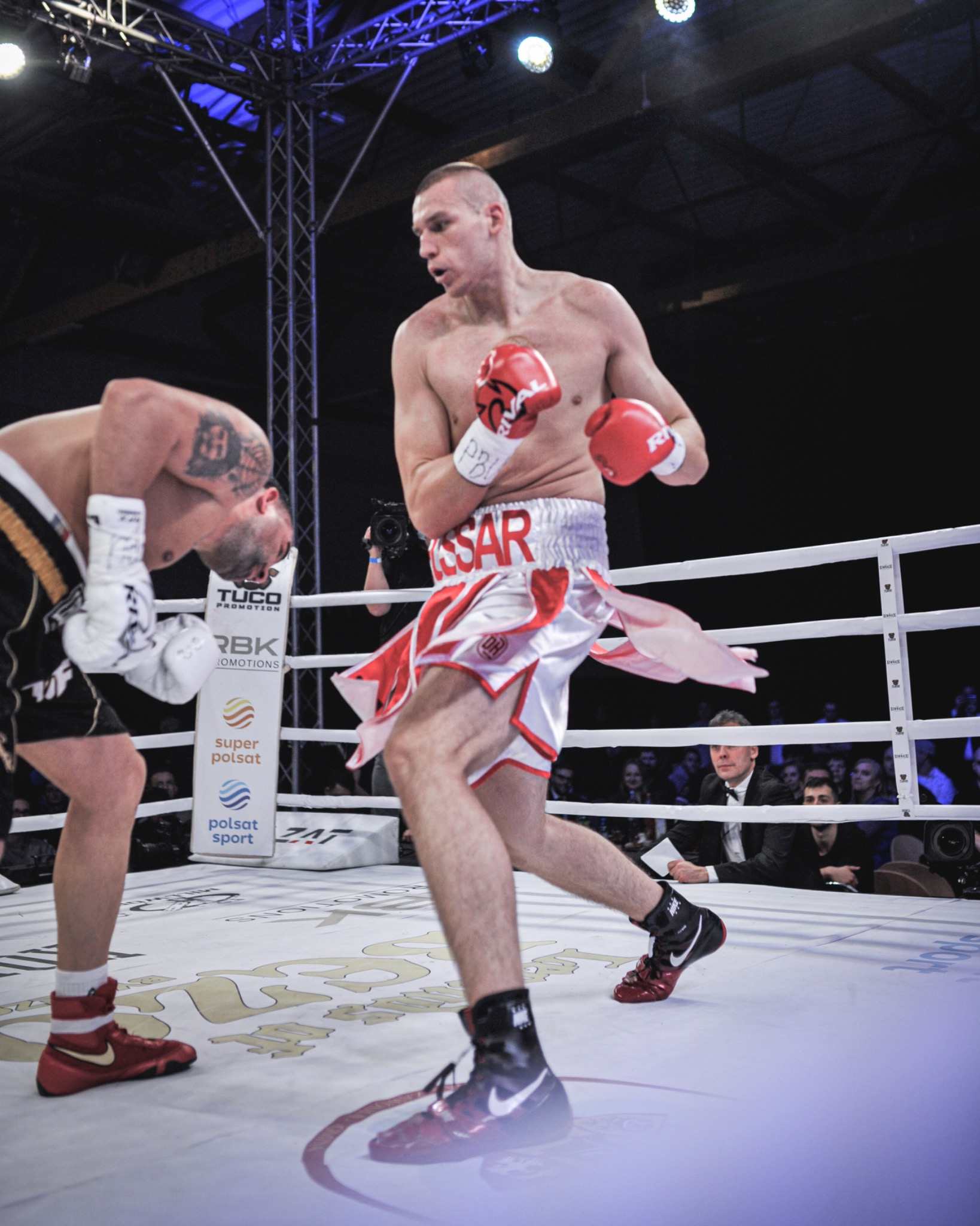 Dynamiczne ujęcie z ringu: bokser w białych spodenkach z czerwonymi akcentami i czerwonych rękawicach w akcji, drugi bokser pochylony, w tle publiczność i oświetlenie hali.