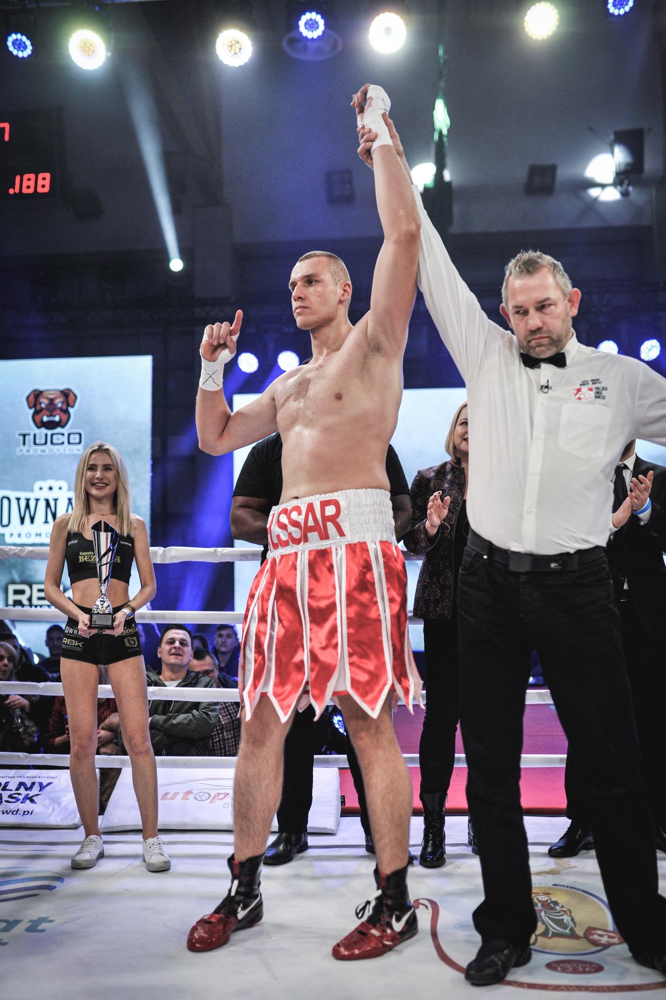 Bokser KSSAR z uniesioną ręką, ogłoszony zwycięzcą walki na ringu. Sędzia w białej koszuli i muszce, ring girl z trofeum, publiczność w tle. Oświetlenie sceniczne.