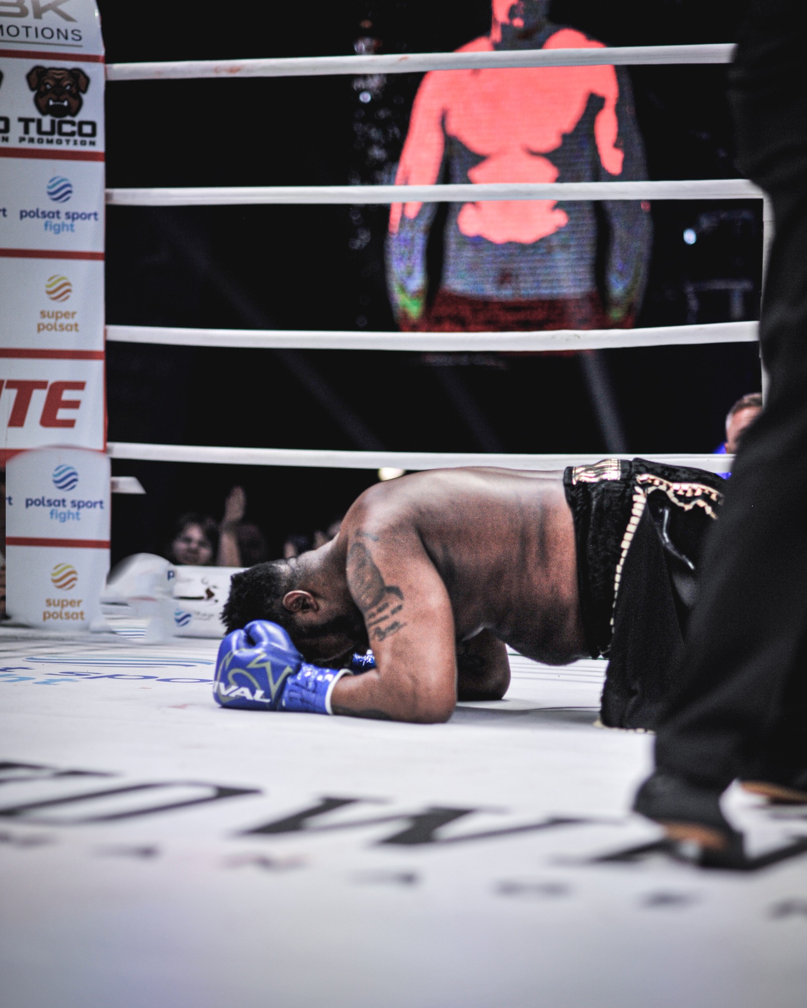 Bokser w niebieskich rękawicach leży na ringu po walce, z widocznymi logotypami sponsorów i publicznością w tle. Ujęcie z bliska, podkreślające emocje.