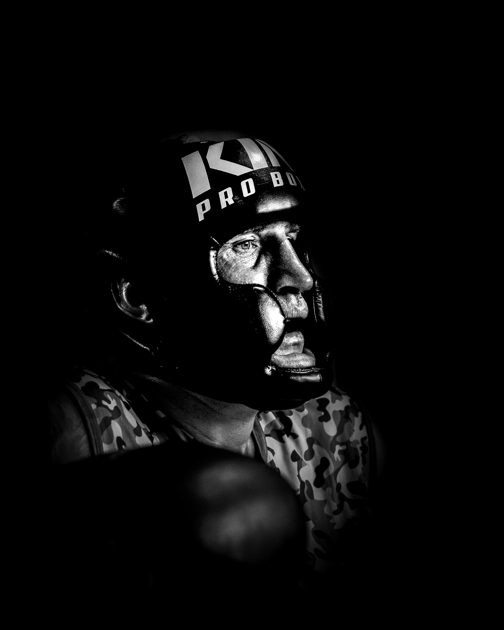 Czarno-biały portret boksera w kasku ochronnym, oświetlonego z boku, z widocznym grymasem na twarzy i koszulką moro. Dynamiczne ujęcie podkreśla determinację sportowca.