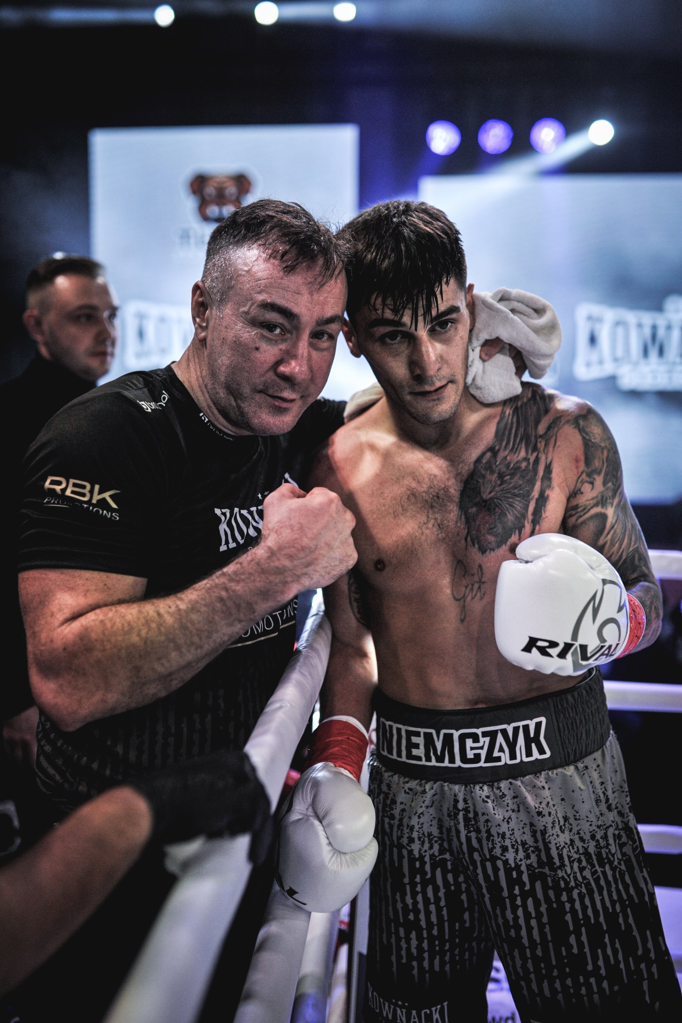 Bokser z trenerem w ringu po walce. Widoczne rękawice, tatuaże i spocone twarze. Oświetlenie sceniczne, dynamiczne ujęcie.