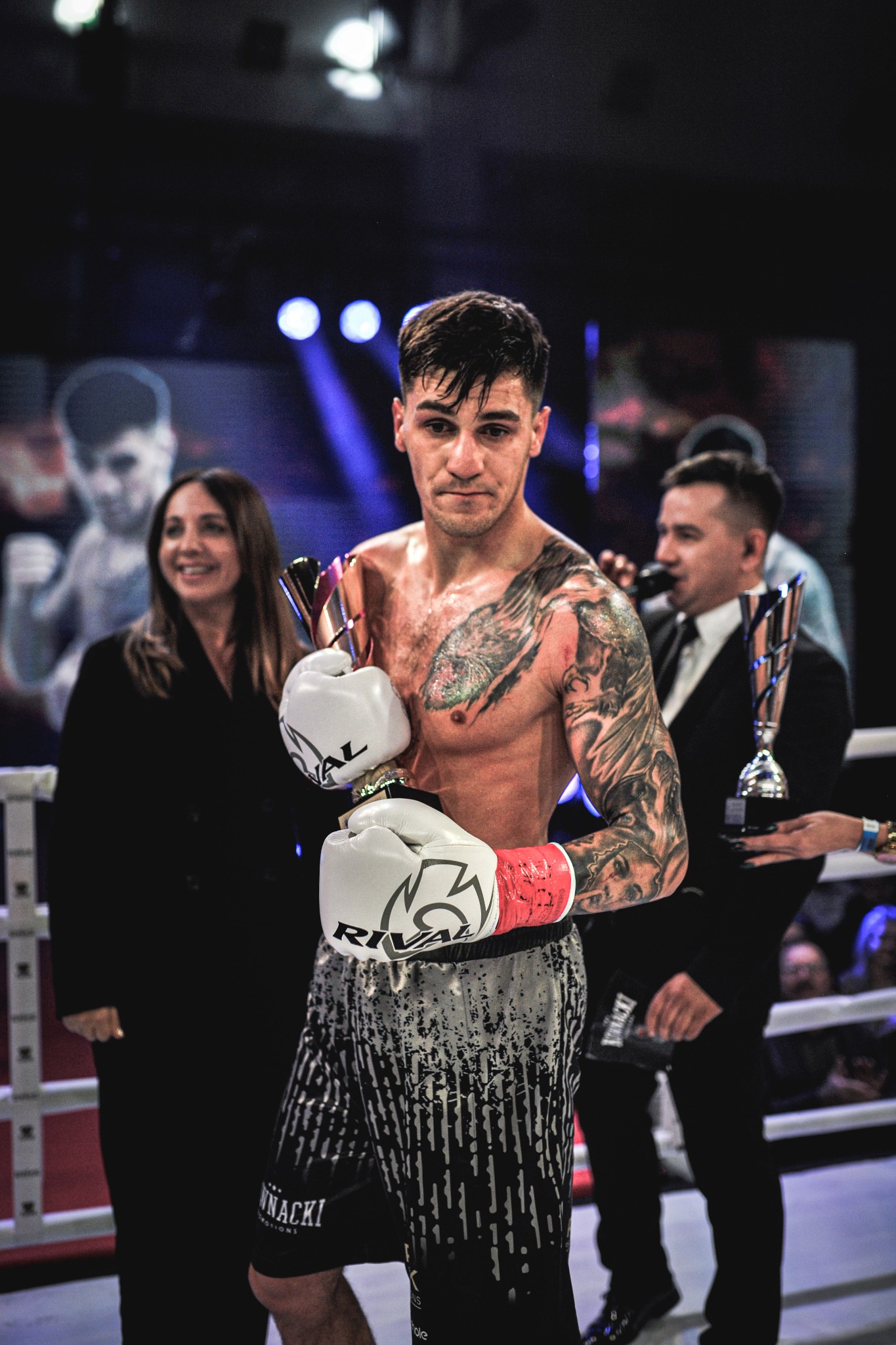 Bokser z tatuażami, w rękawicach Rival, trzyma trofeum na ringu. W tle kobieta i mężczyzna z mikrofonem. Scena po walce, oświetlenie sceniczne.