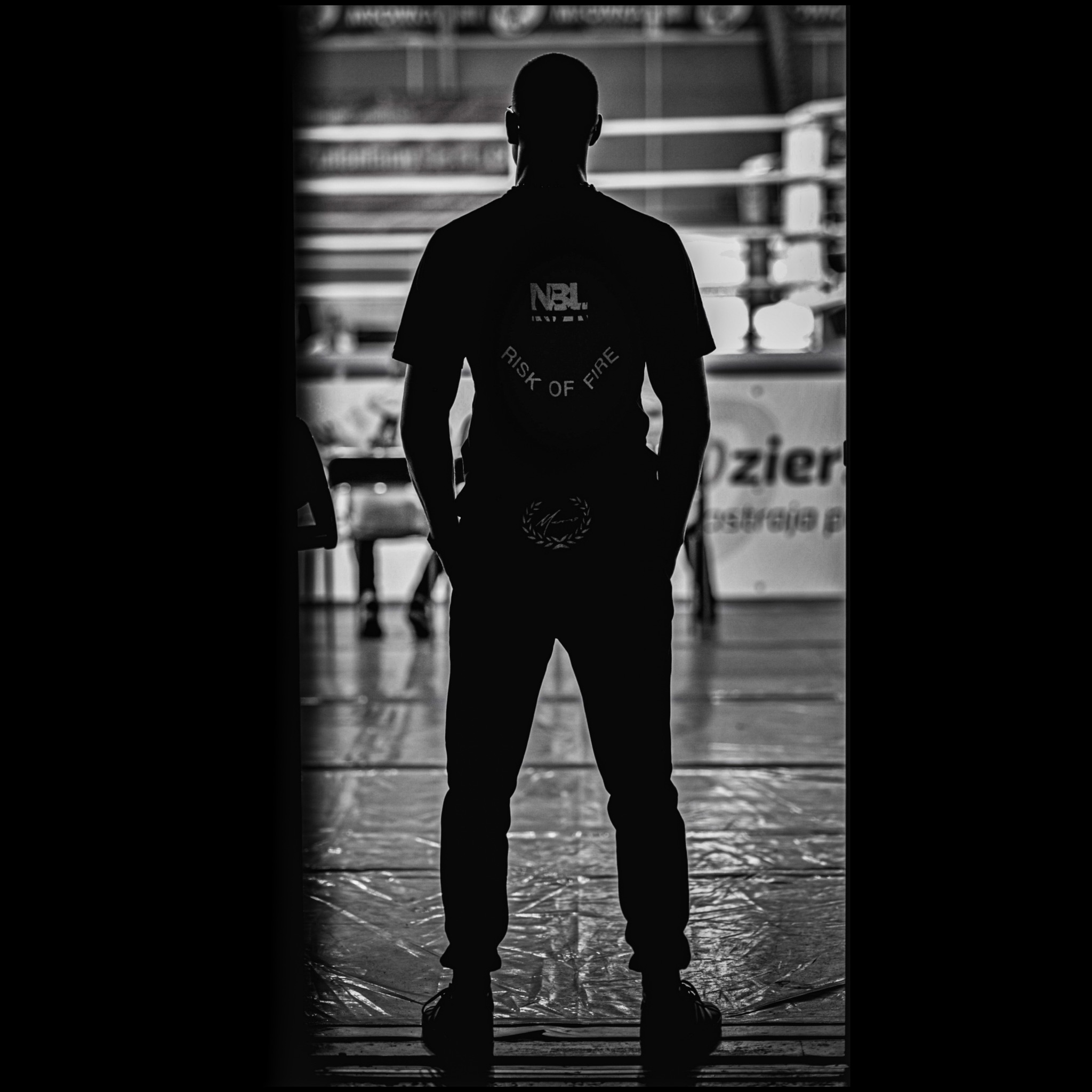 Sylwetka mężczyzny w czarnej koszulce z logo NBL, stojącego tyłem na tle ringu bokserskiego. Monochromatyczne ujęcie z ciemnymi krawędziami.