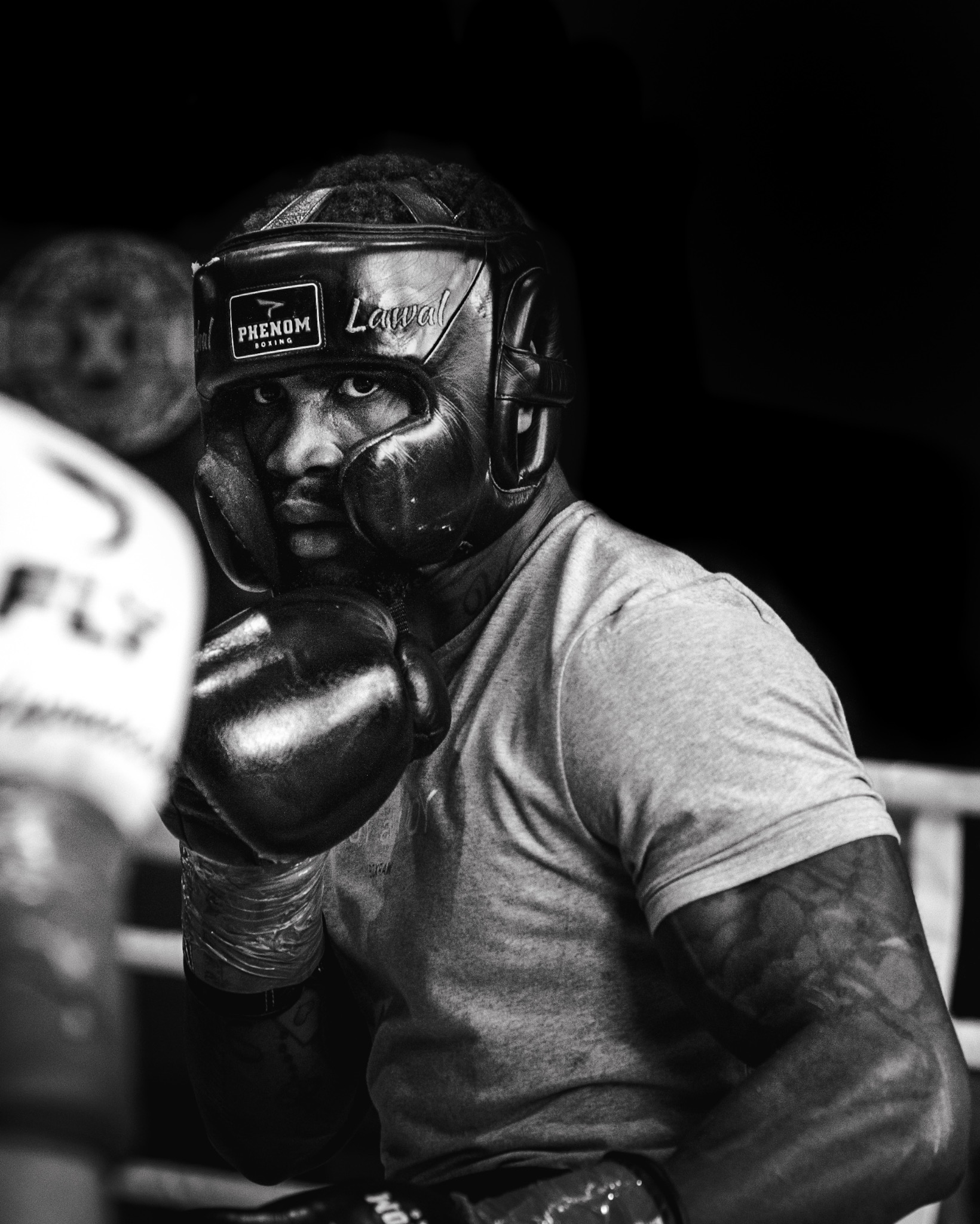 Czarno-białe zdjęcie boksera w ringu, w kasku i rękawicach, z intensywnym spojrzeniem. Widoczne detale: nazwa marki na kasku, bandaże na rękach, tatuaże na ramieniu.