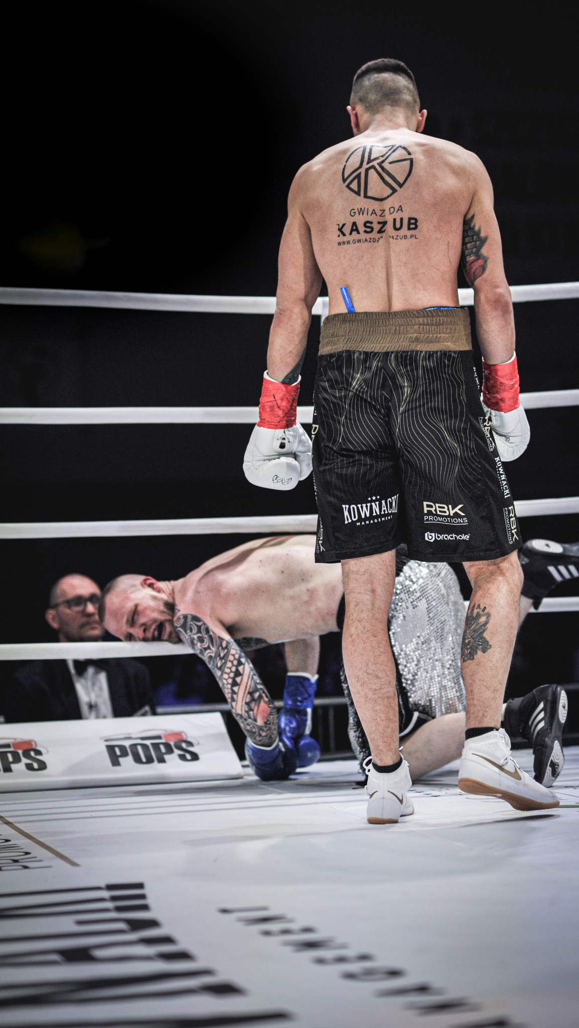 Widok z tyłu na boksera z tatuażem na plecach, stojącego nad powalonym przeciwnikiem na ringu. W tle widoczni widzowie. Dynamiczne ujęcie z gali bokserskiej.