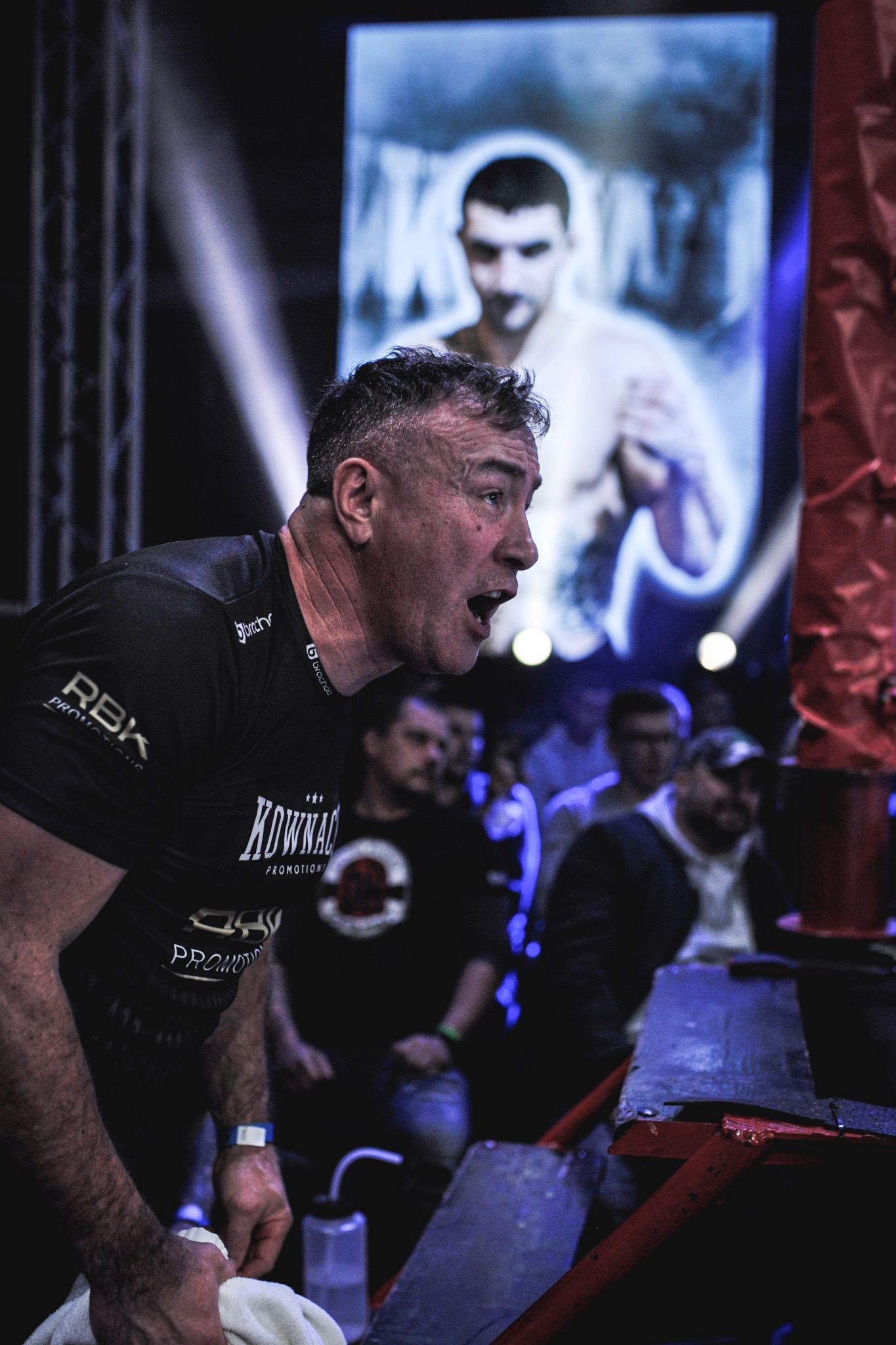 Trener boksu krzyczy, trzymając ręcznik, w tle ekran z wizerunkiem boksera. Dynamiczne ujęcie z ringu podczas wydarzenia sportowego, oświetlenie sceniczne.