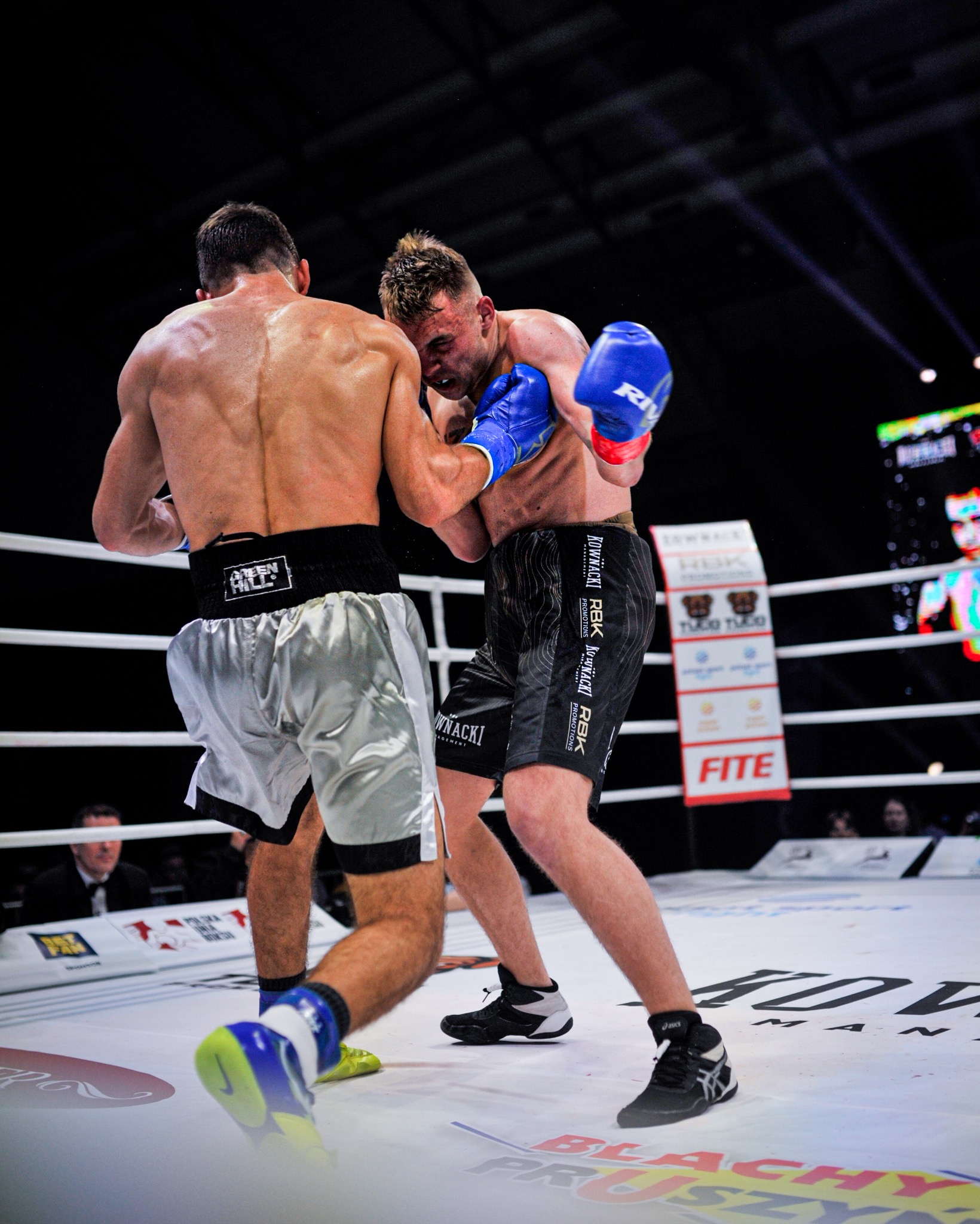 Dynamiczne ujęcie z ringu: dwóch bokserów w zwarciu, jeden z niebieskimi rękawicami zadaje cios. Widoczne logo sponsorów i oświetlenie hali.