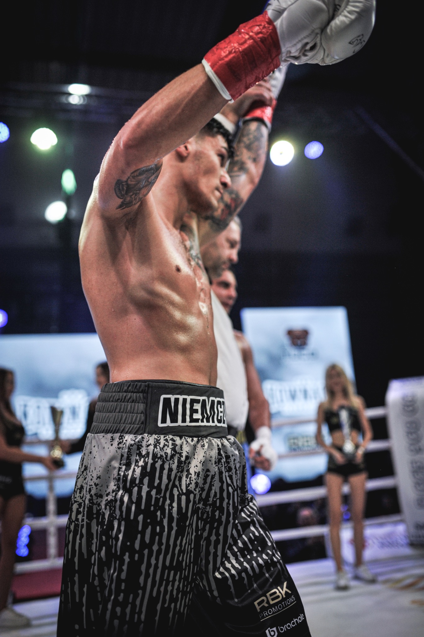 Zwycięski bokser z podniesionymi rękami w ringu, w tle rozmazane postacie i światła. Na spodenkach widoczny napis NIEMIEC i logo RBK Promotions.