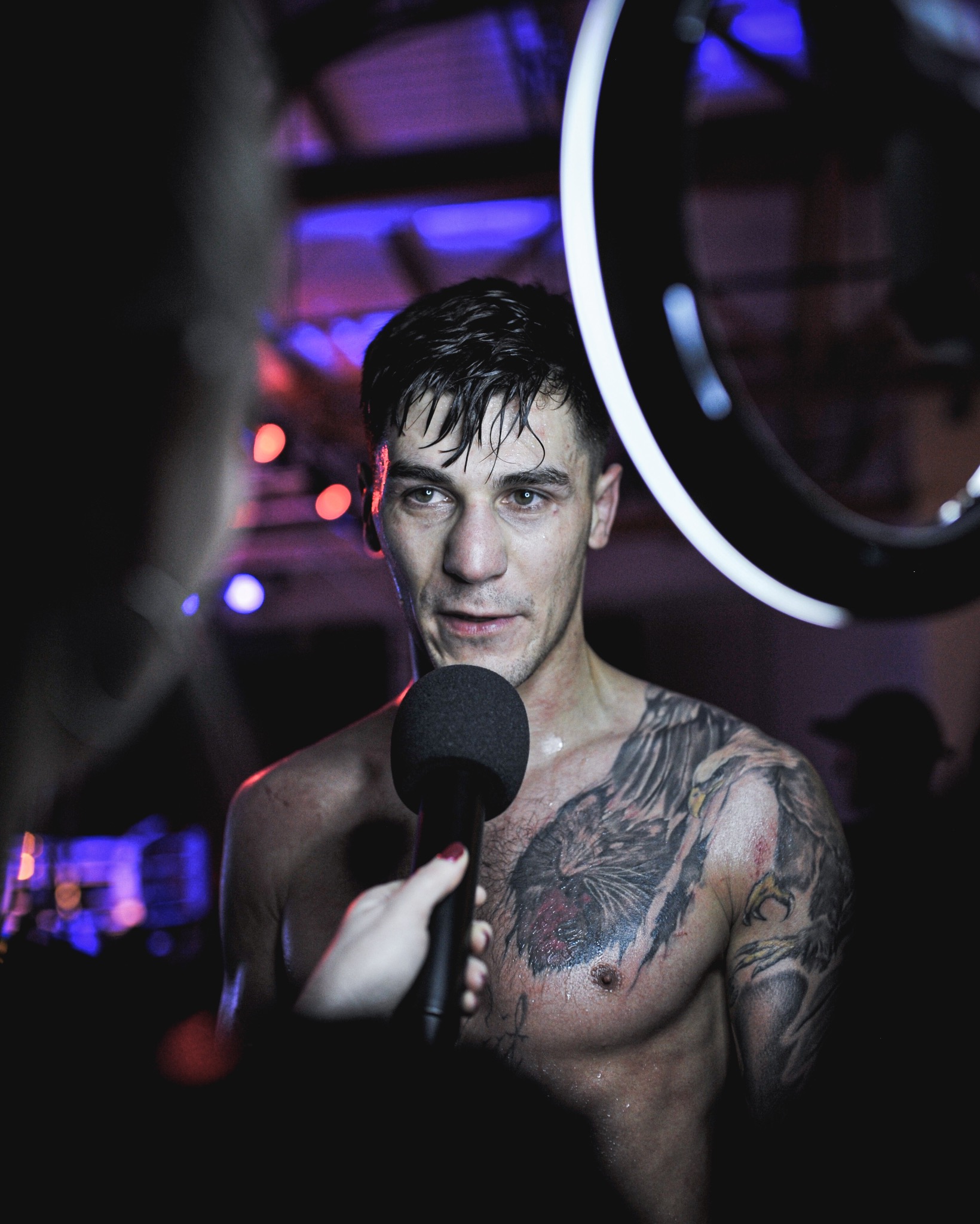 Mężczyzna z tatuażem na ramieniu, spocony, trzyma mikrofon podczas wywiadu na tle sceny z oświetleniem. Ujęcie z bliska, dynamiczne światło.