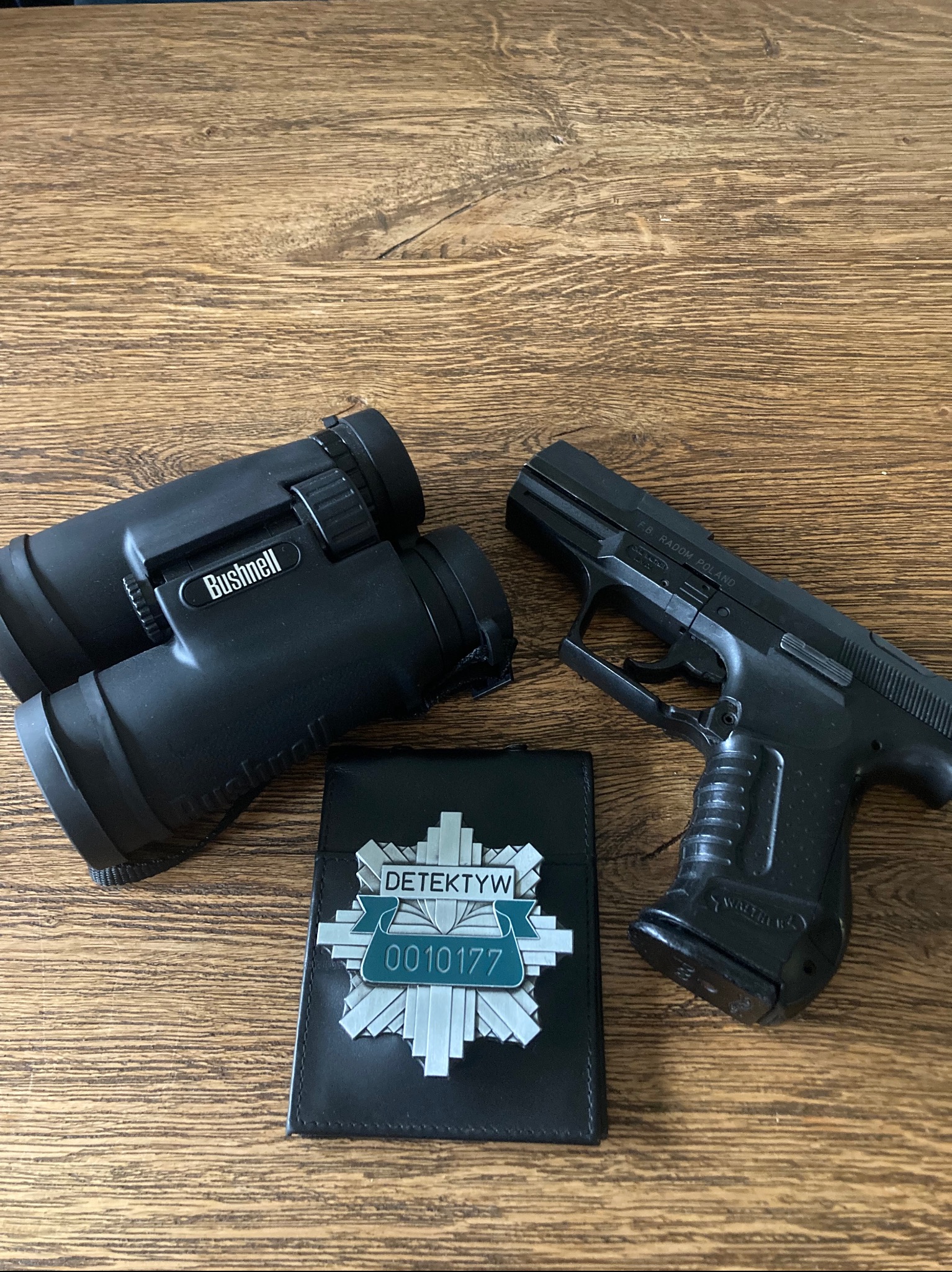 Czarny portfel z odznaką 'Detektyw', lornetka Bushnell i pistolet Walther na drewnianym stole. Sprzęt detektywistyczny w zbliżeniu.