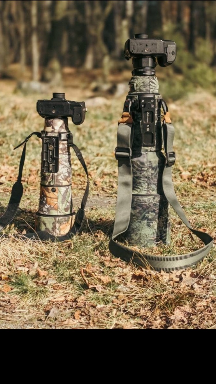 Dwa aparaty fotograficzne z teleobiektywami w kamuflażu, stojące na trawie w plenerze. Jeden obiektyw z szerszym paskiem, drugi z węższym. Tło rozmyte, leśne.