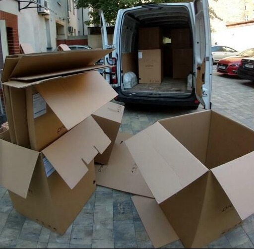 Kartonowe pudła przed busem z otwartymi drzwiami, gotowe do załadunku. Część pudeł leży na ziemi, inne są otwarte i czekają na wypełnienie.