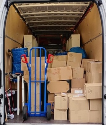 Wnętrze busa wypełnione kartonami i wózkiem transportowym z niebieską ramą i czerwonymi uchwytami, gotowe do transportu. Widoczne torby IKEA.