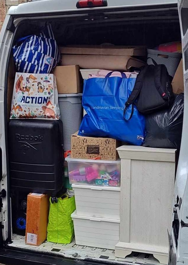 Wnętrze busa wypełnione po brzegi rzeczami do przeprowadzki: torby, kartony, walizka Reebok, meble. Widok z tyłu pojazdu.