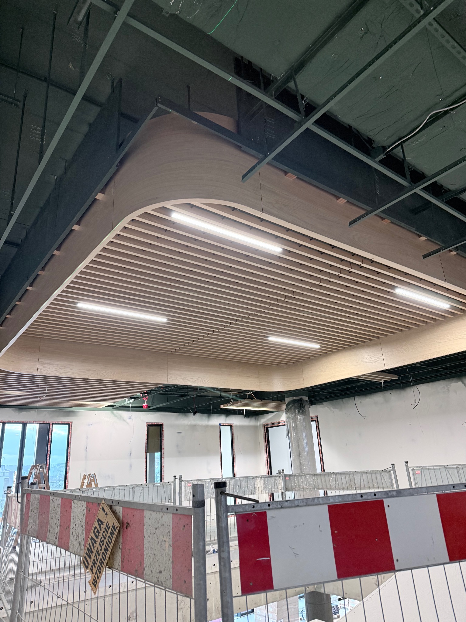 Drewniana, podwieszana konstrukcja sufitowa z oświetleniem liniowym w biurze, widoczna z dołu. W tle surowy stan ścian i zabezpieczenia budowlane.