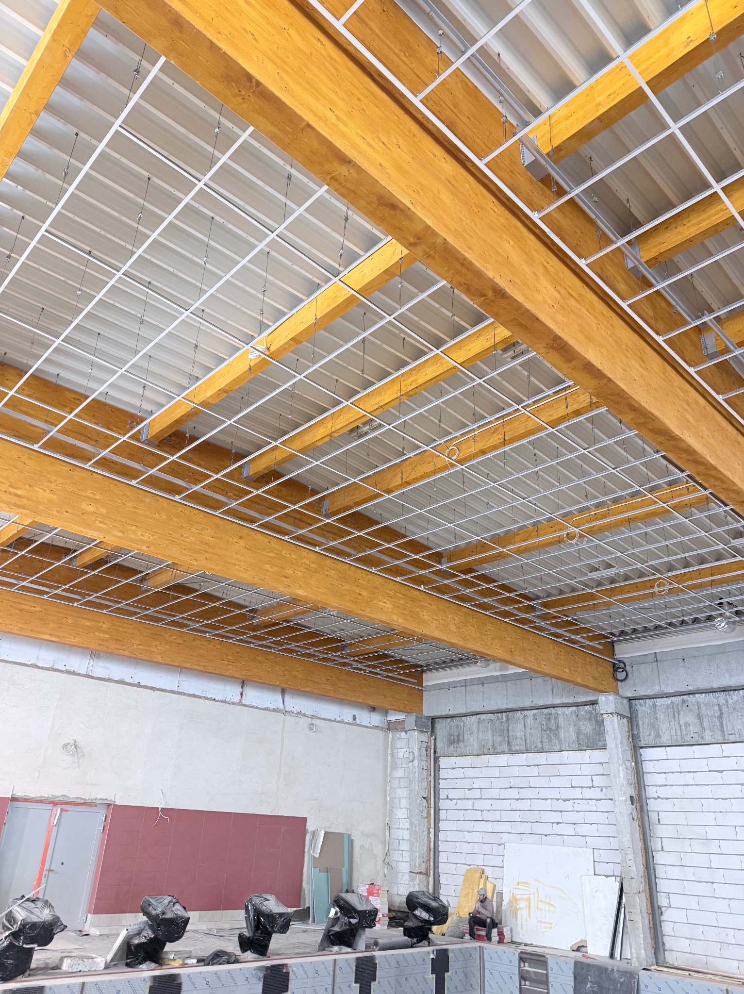 Montaż sufitu podwieszanego z drewnianymi belkami i metalową konstrukcją w przestrzeni komercyjnej. Widoczne elementy instalacji i surowy stan ścian.