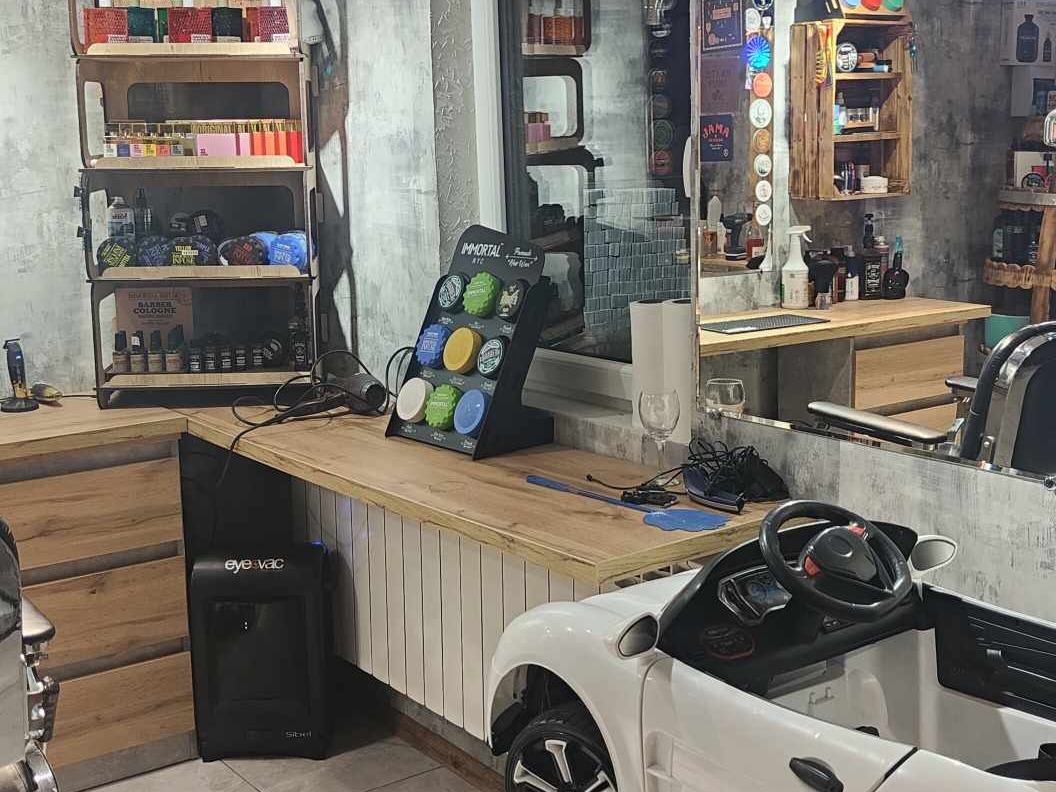 Wnętrze salonu barberskiego z drewnianymi półkami, kosmetykami, lustrem i dziecięcym autkiem, tworzące unikalny, przyjazny klimat.