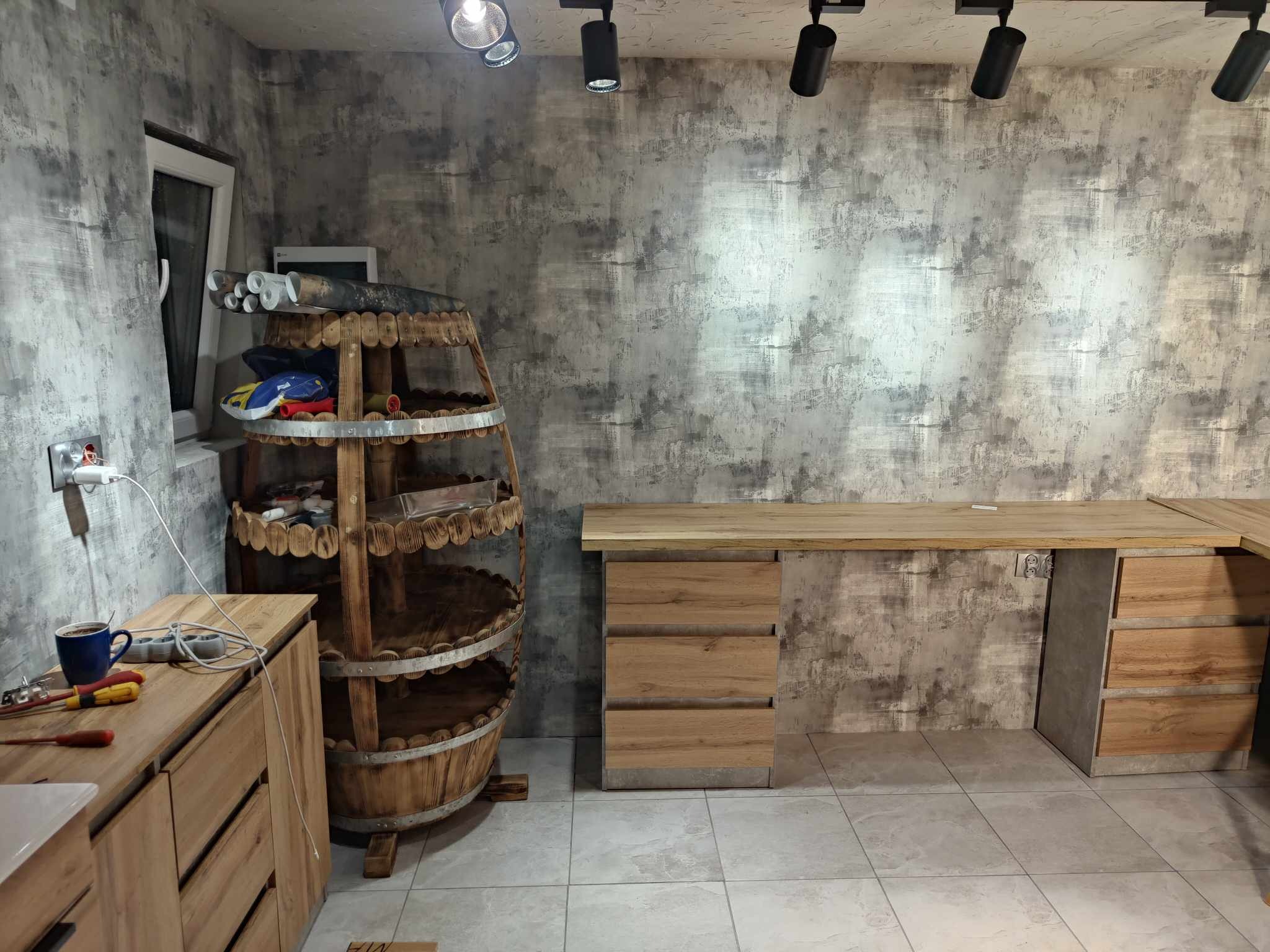 Drewniane meble z szufladami i biurko w pomieszczeniu z szarą ścianą. Dekoracyjny regał w kształcie beczki. Widoczne narzędzia i akcesoria.
