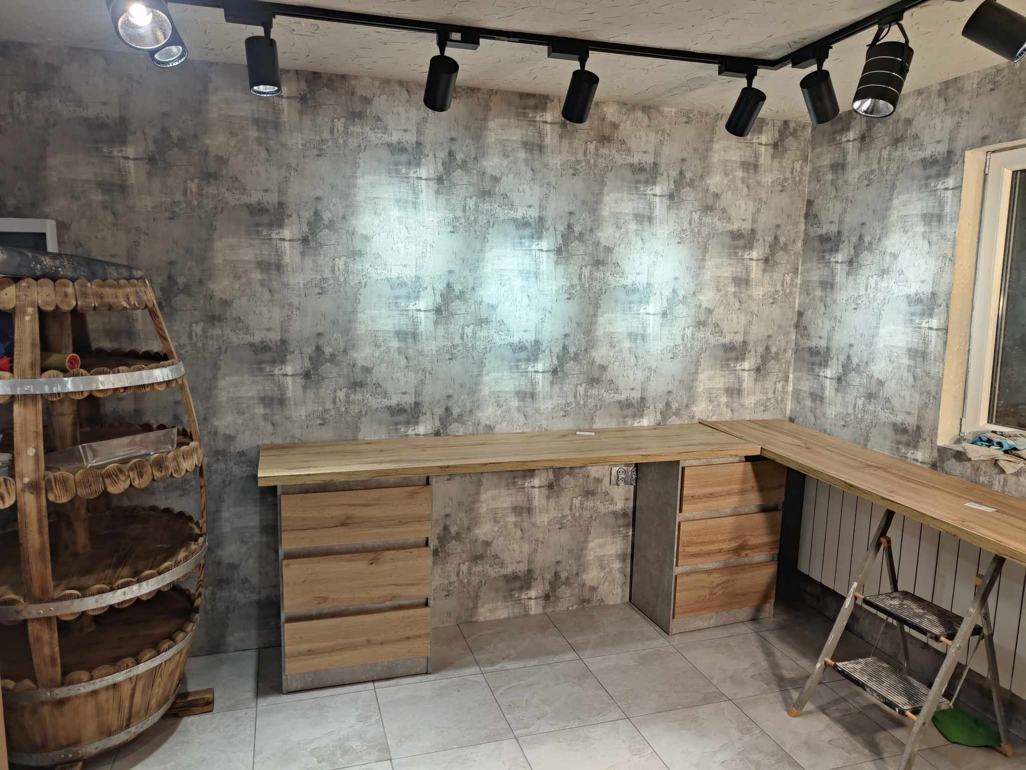 Nowoczesne biuro z drewnianym blatem i szufladami, szara ściana z fakturą, oświetlenie punktowe i dekoracyjna beczka. Widoczna drabina.